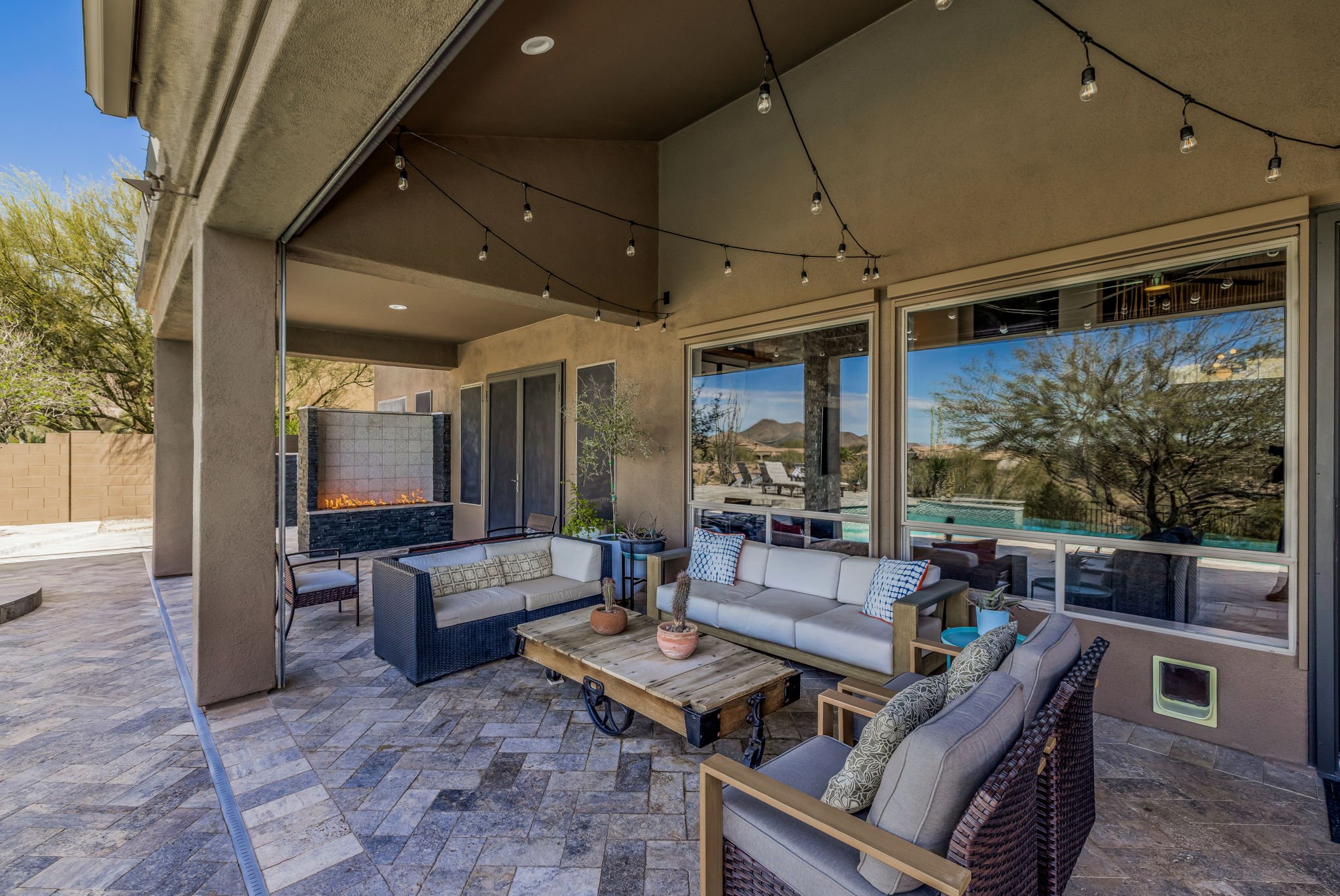Covered outdoor patio with seating area including a white sofa, armchair, and a rustic wooden coffee table, string lights hanging above, fireplace on wall, large windows reflecting desert landscape with mountains