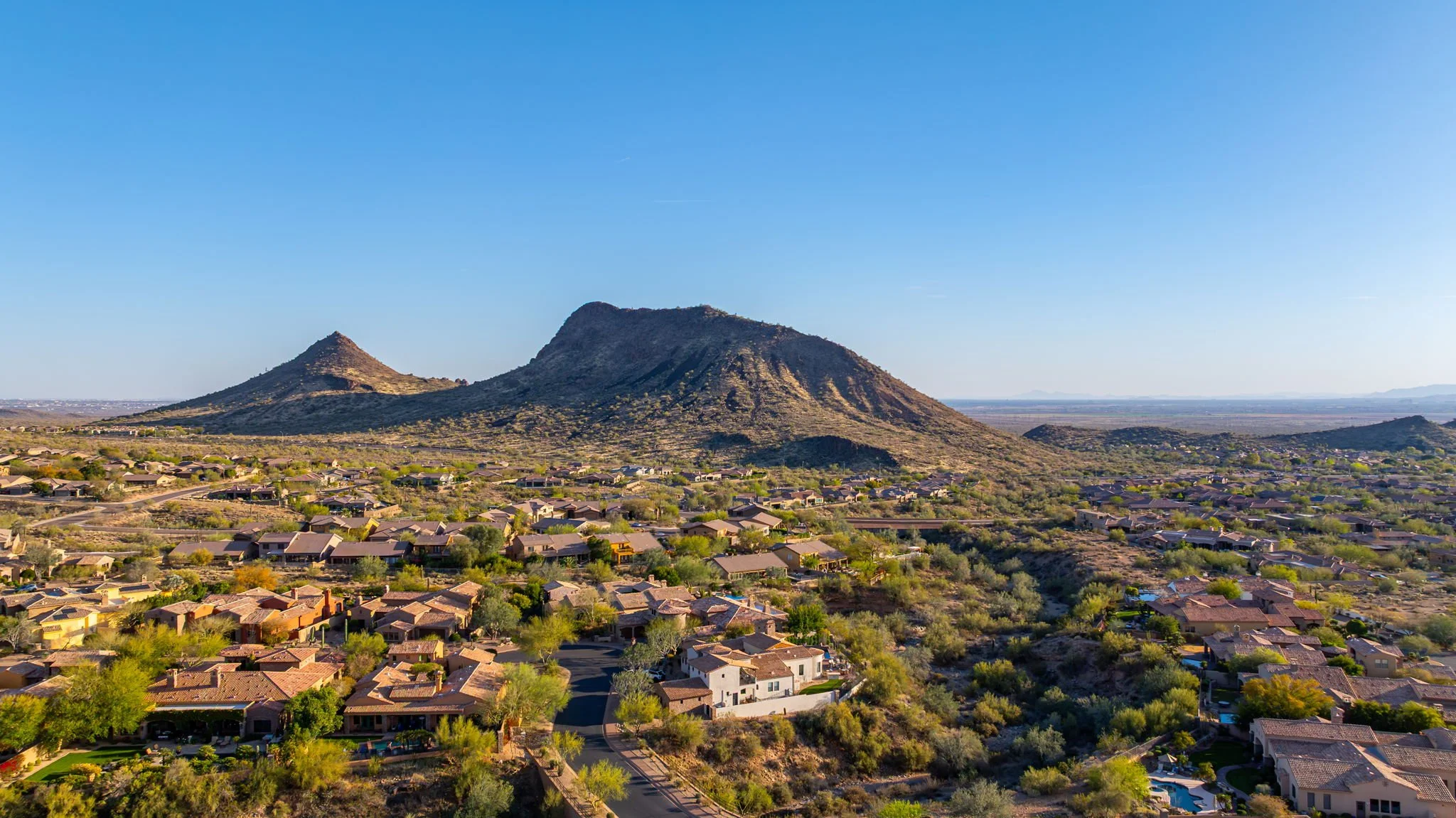 Aerial view of a residential neighborhood with desert houses and lush trees, set against a backdrop of mountains and a clear blue sky.