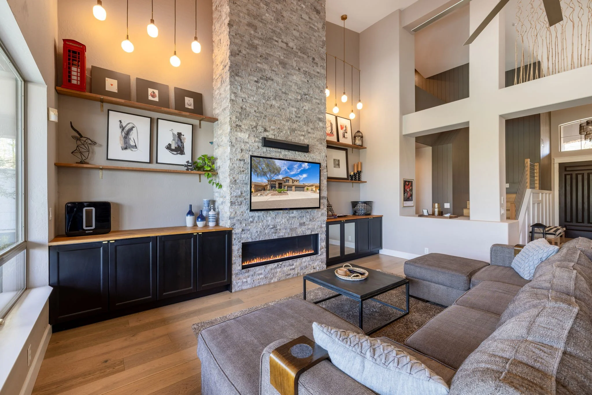 Modern living room with a sectional sofa, media wall with a TV above an electric fireplace, floating shelves with artwork, decorative items, and a view of a staircase and front entrance in the background.