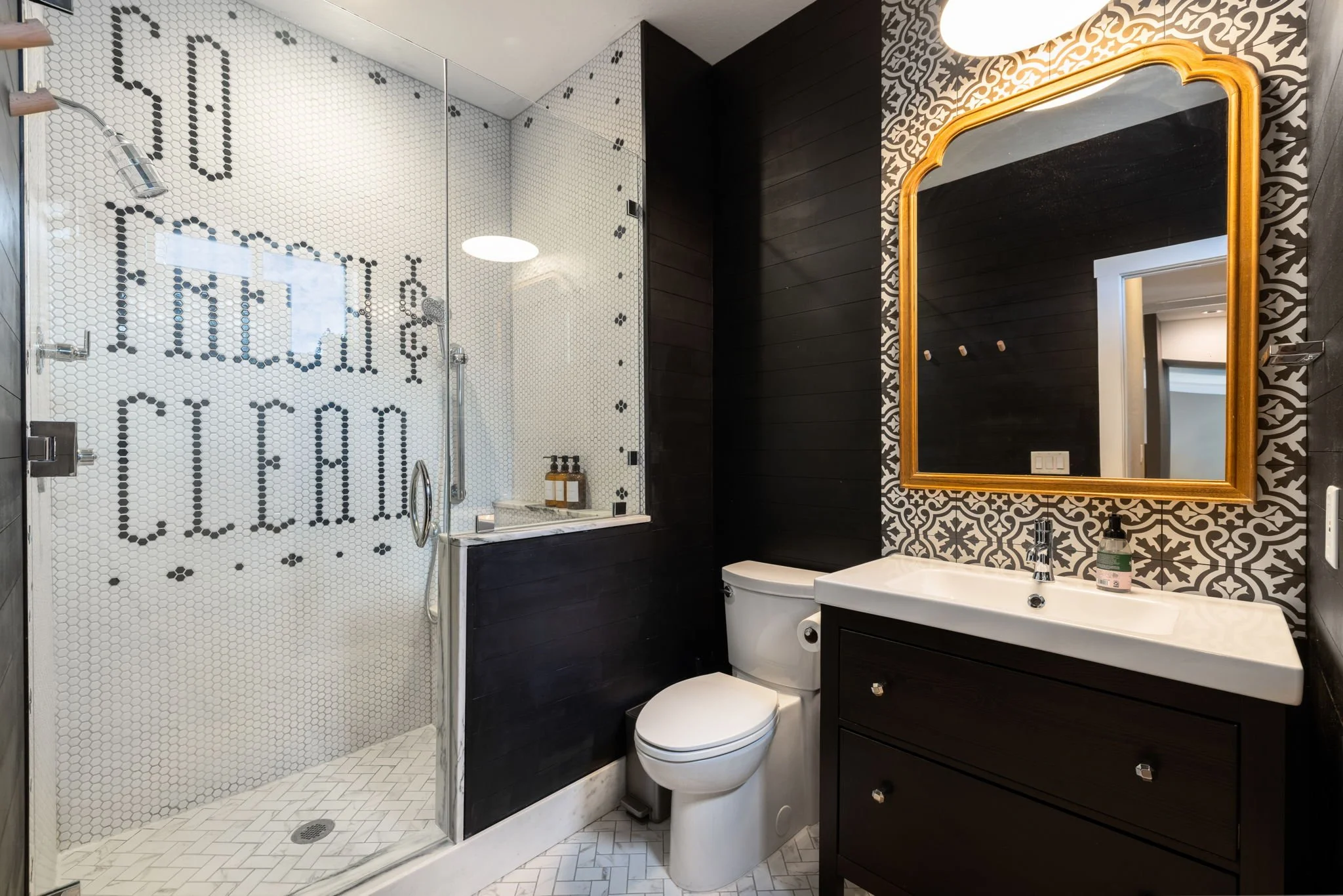 Modern bathroom with a walk-in shower featuring a glass door and a mosaic wall with the words 'So Fresh, So Clean' in black tiles. There is a white toilet and a black vanity with a white sink and a large mirror with a gold frame. The wall behind the 