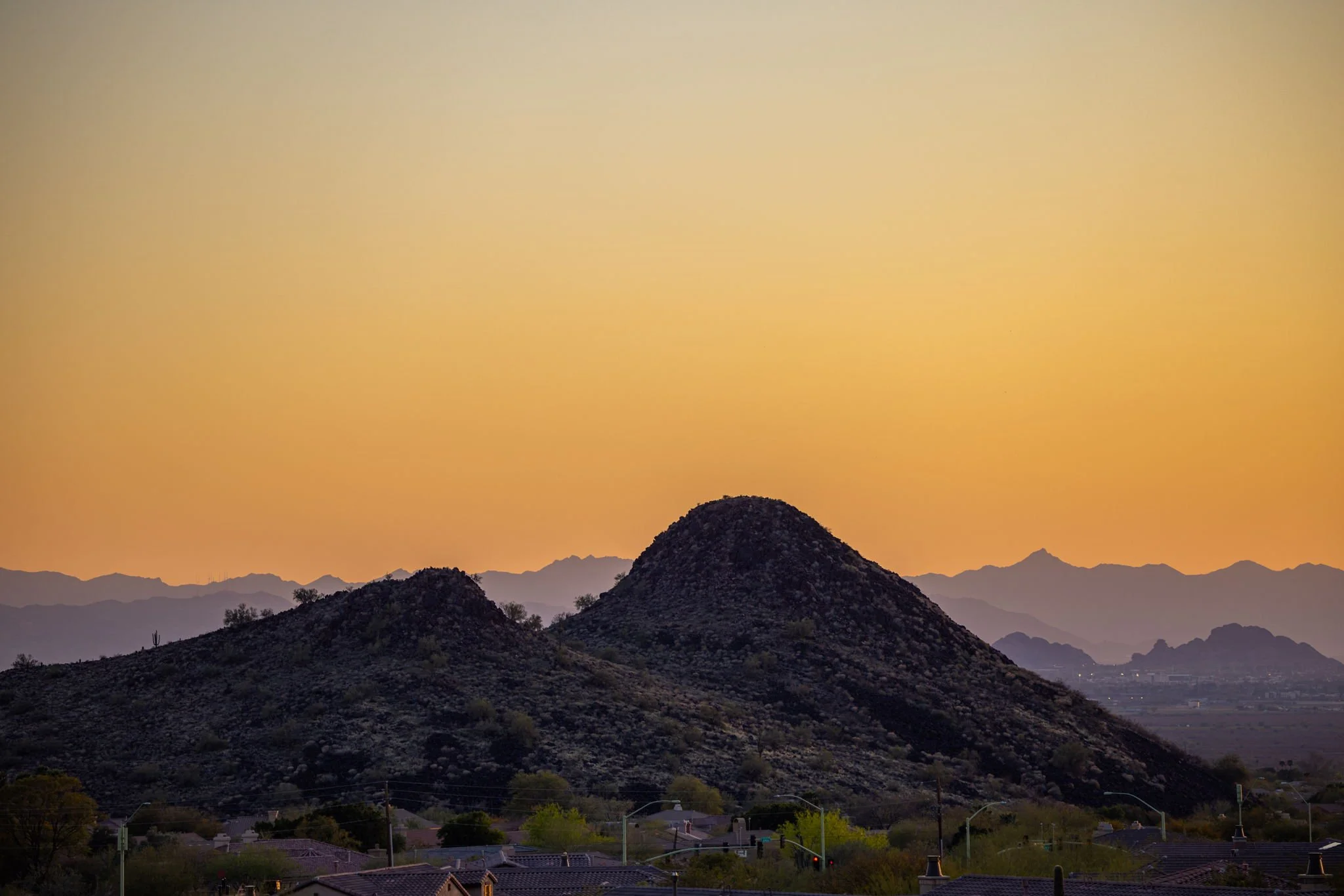 Silhouetted mountains at sunset with a dull orange sky, view from a suburban area with rooftops and trees in the foreground.