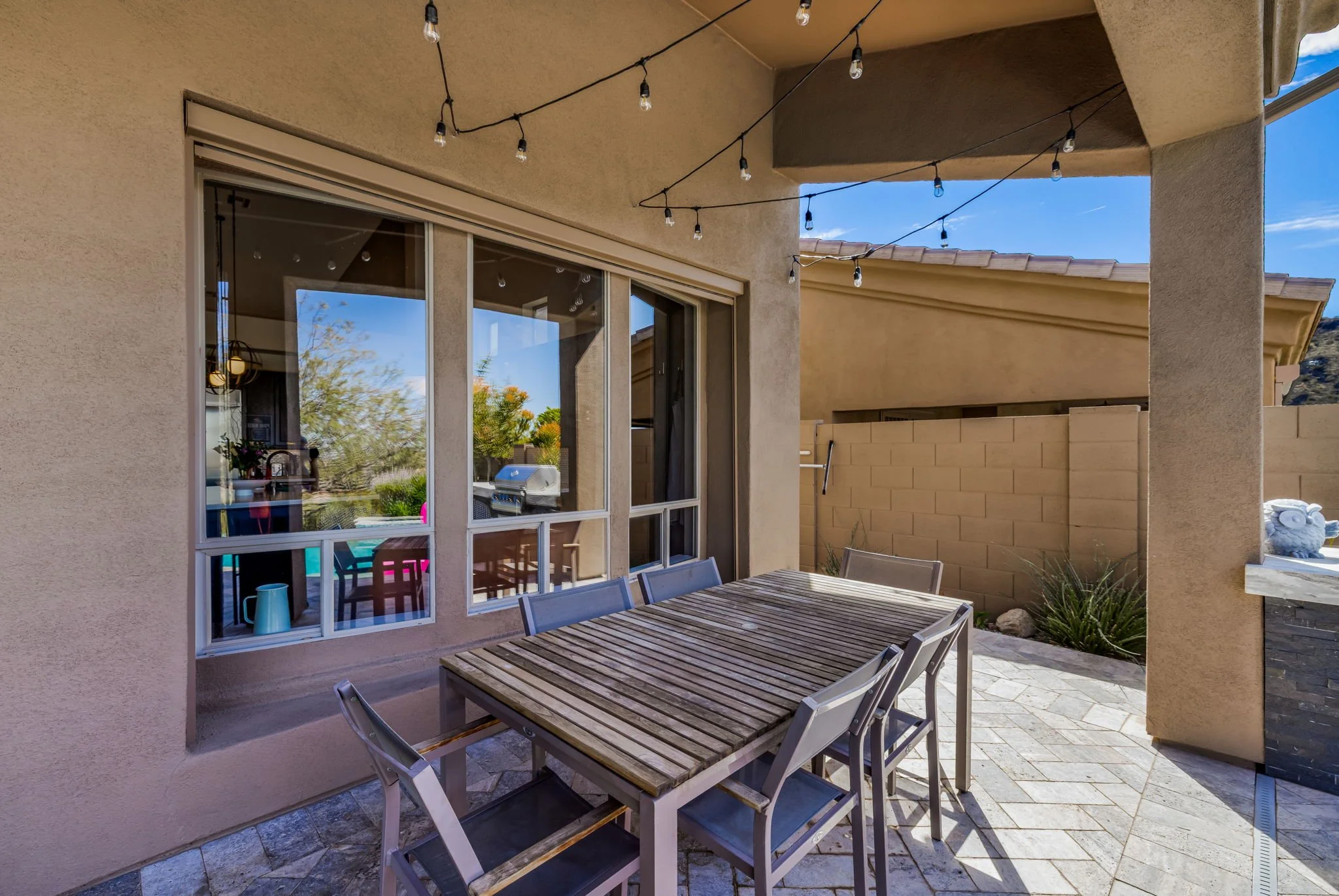 Outdoor patio with a wooden dining table and six metal chairs, string lights overhead, beige stucco walls, and a large window reflecting a blue sky with clouds and some trees.