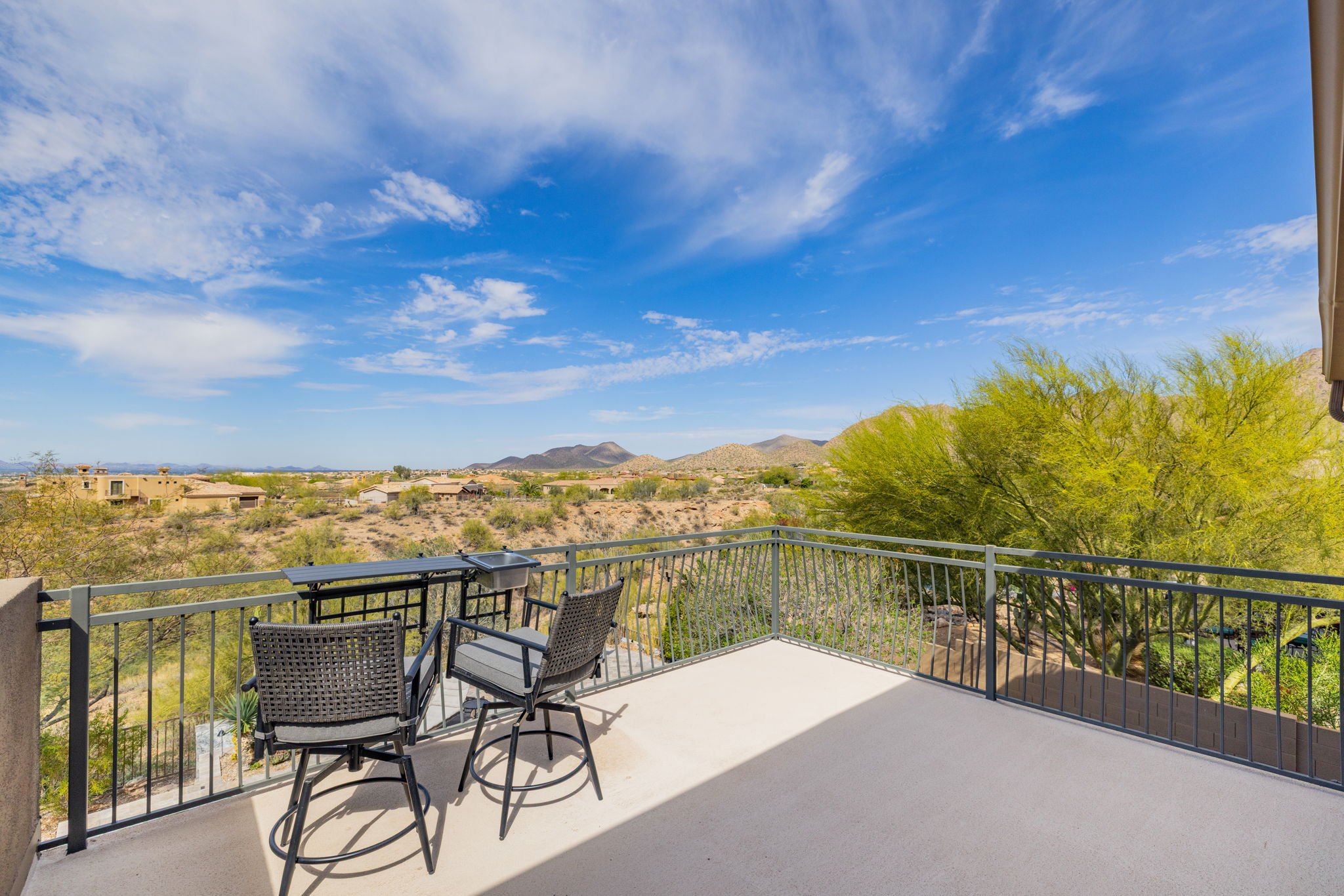 View from a balcony overlooking a desert landscape with mountains, houses, a green tree, and a blue sky with clouds.