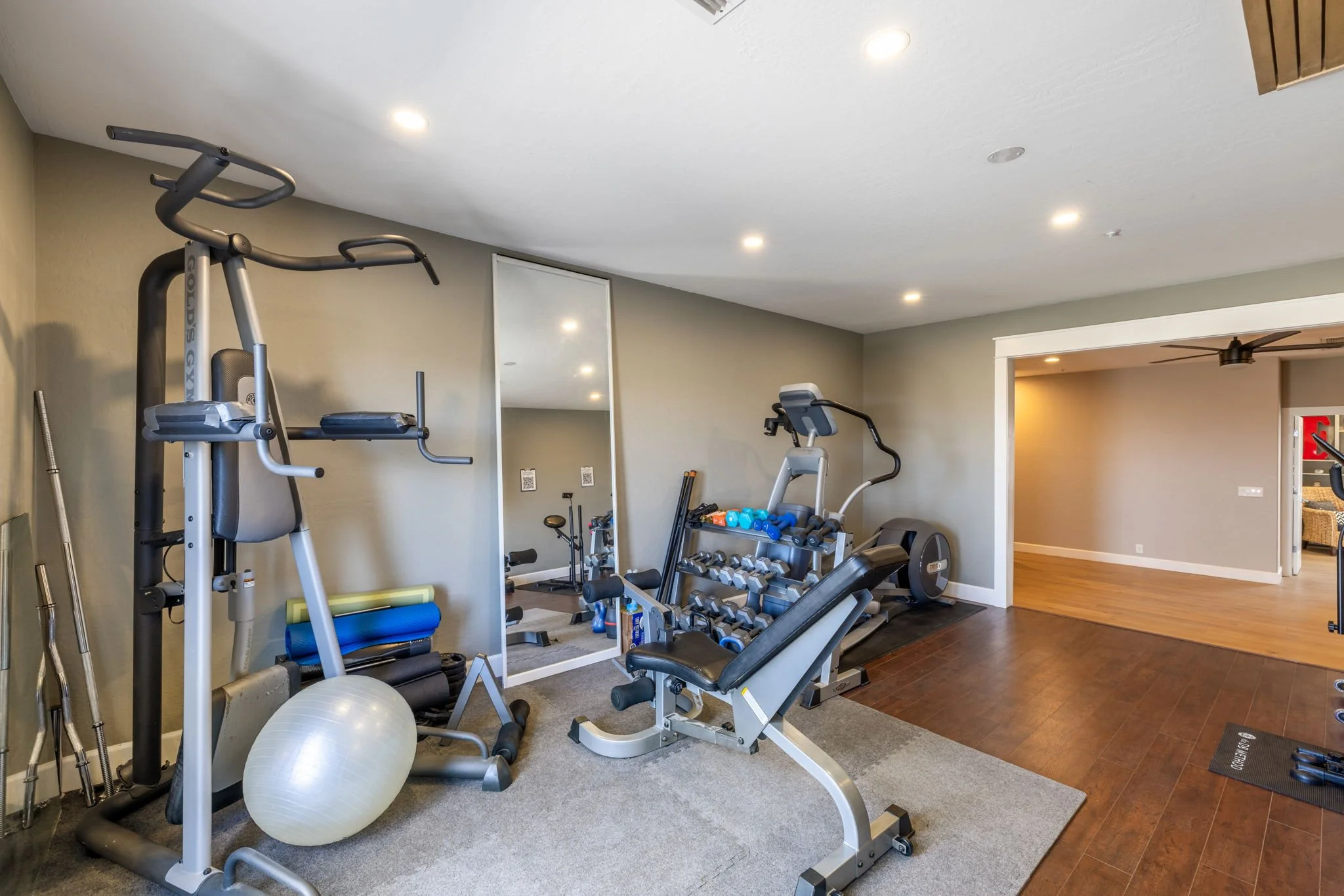 Home gym with exercise equipment including a weight machine, dumbbells on a rack, additional cardio equipment, a stability ball, and a mirror on the wall. The room has recessed lighting and hardwood flooring.