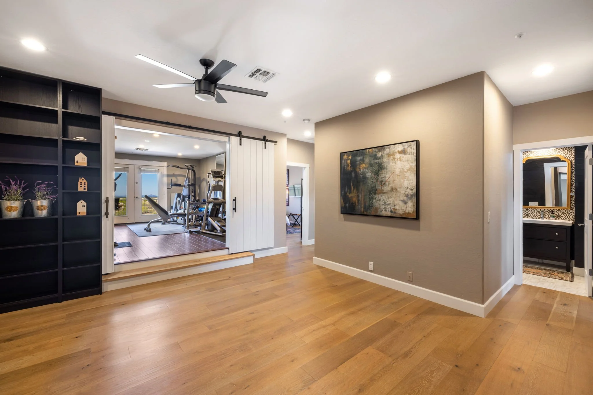 Living room with wooden floors, a black ceiling fan, beige walls, and artwork on the wall. An open doorway with black framing leads to a home gym with workout equipment and sliding barn doors.
