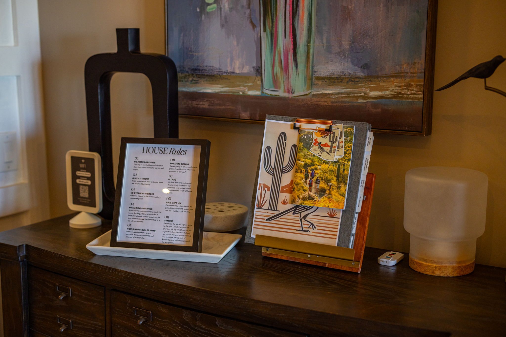 A wooden sideboard with a framed house rules sheet, a brochure stand with desert-themed images and postcards, a modern white table lamp with a cream-colored shade, a small QR code display, and a small white remote control. There is a large abstract p