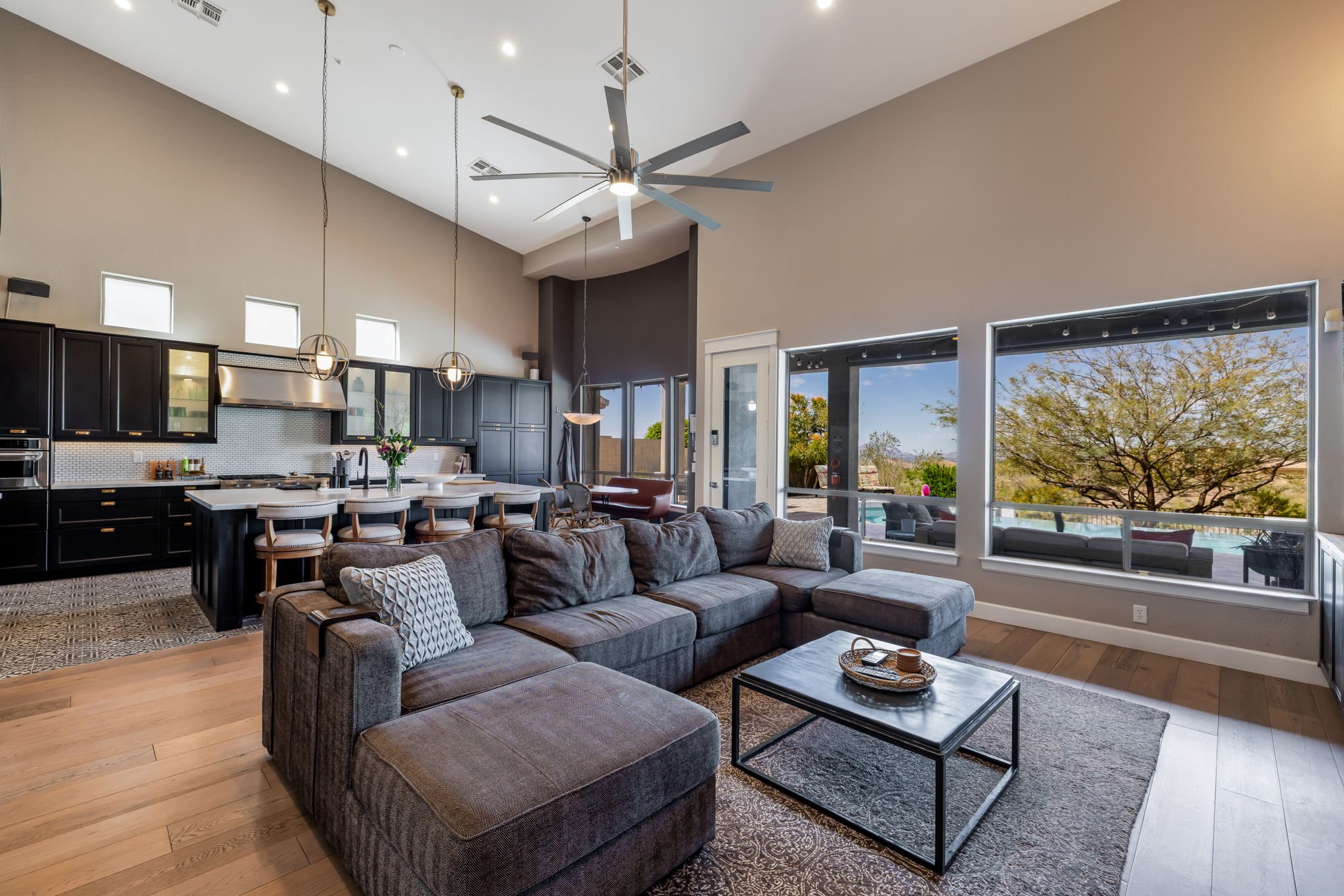 Open-concept living room and kitchen with large windows, gray sectional sofa, black kitchen cabinets, island with bar stools, and a view of an outdoor pool and trees.