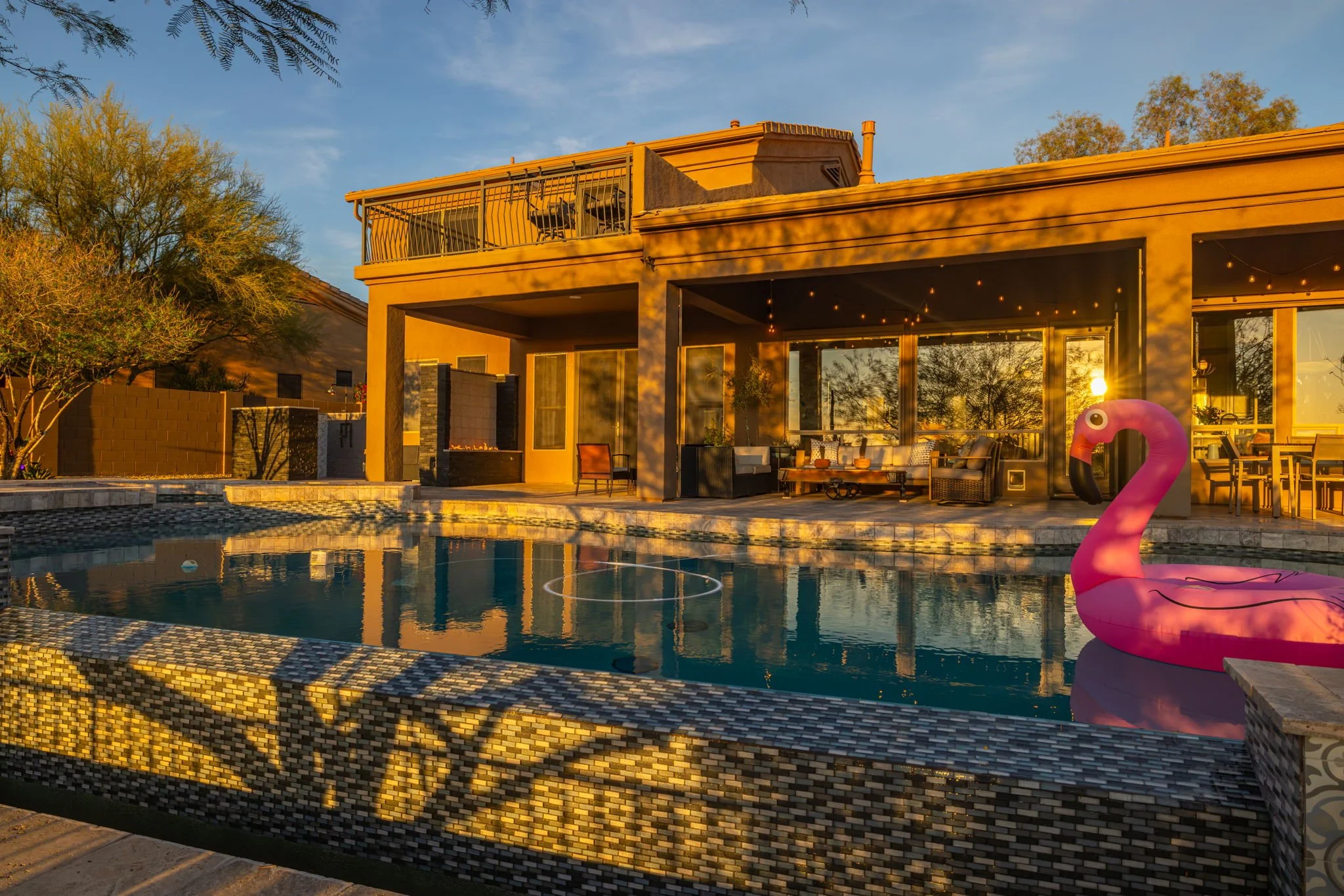 A backyard with a swimming pool and an inflatable pink flamingo in the water. There is a modern house with large windows and outdoor furniture, and a tree on the left side. The scene is during sunset or sunrise, with warm lighting.