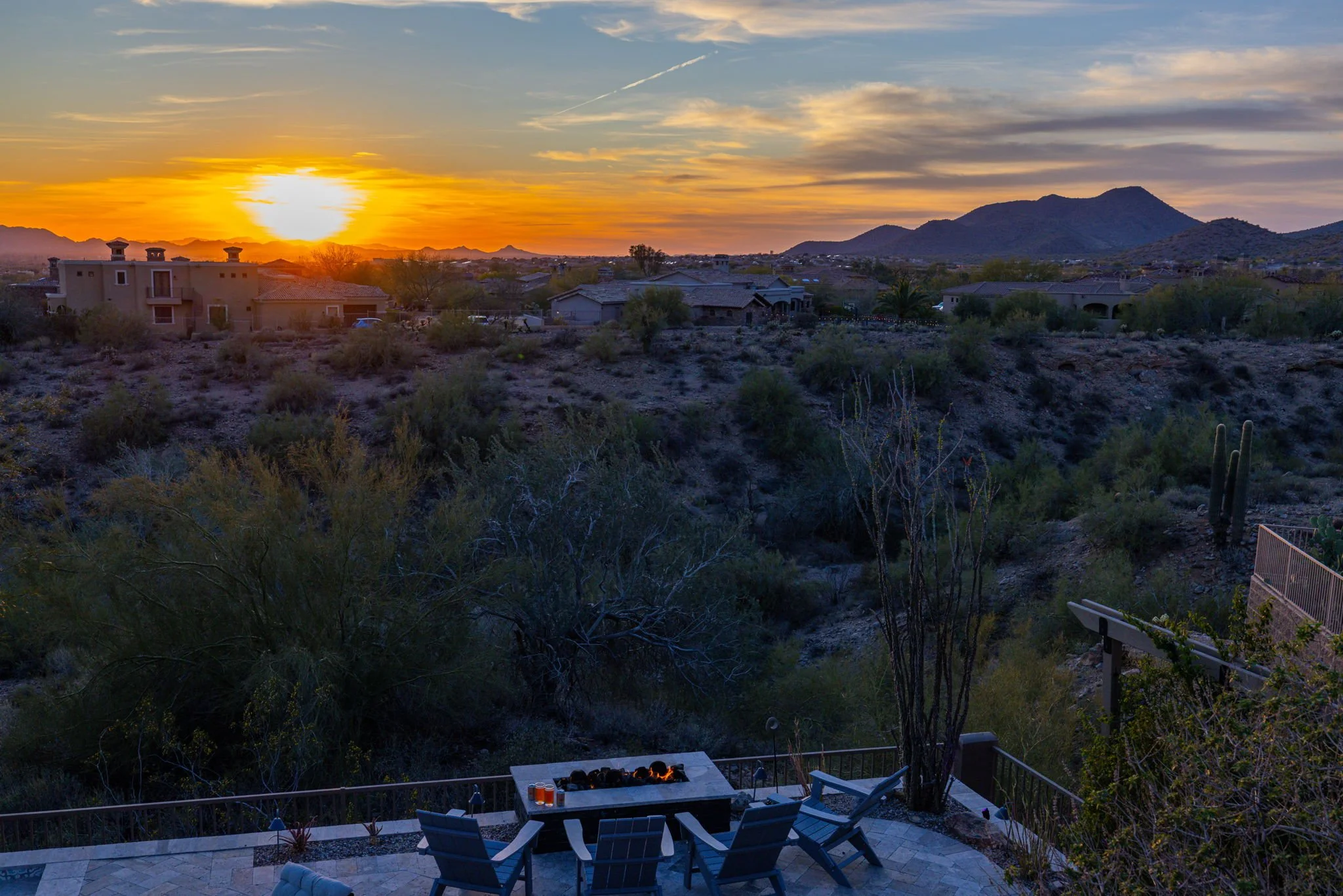 Sunset over a desert landscape with houses, mountains, and sparse vegetation, viewed from a patio with outdoor chairs and a firepit.