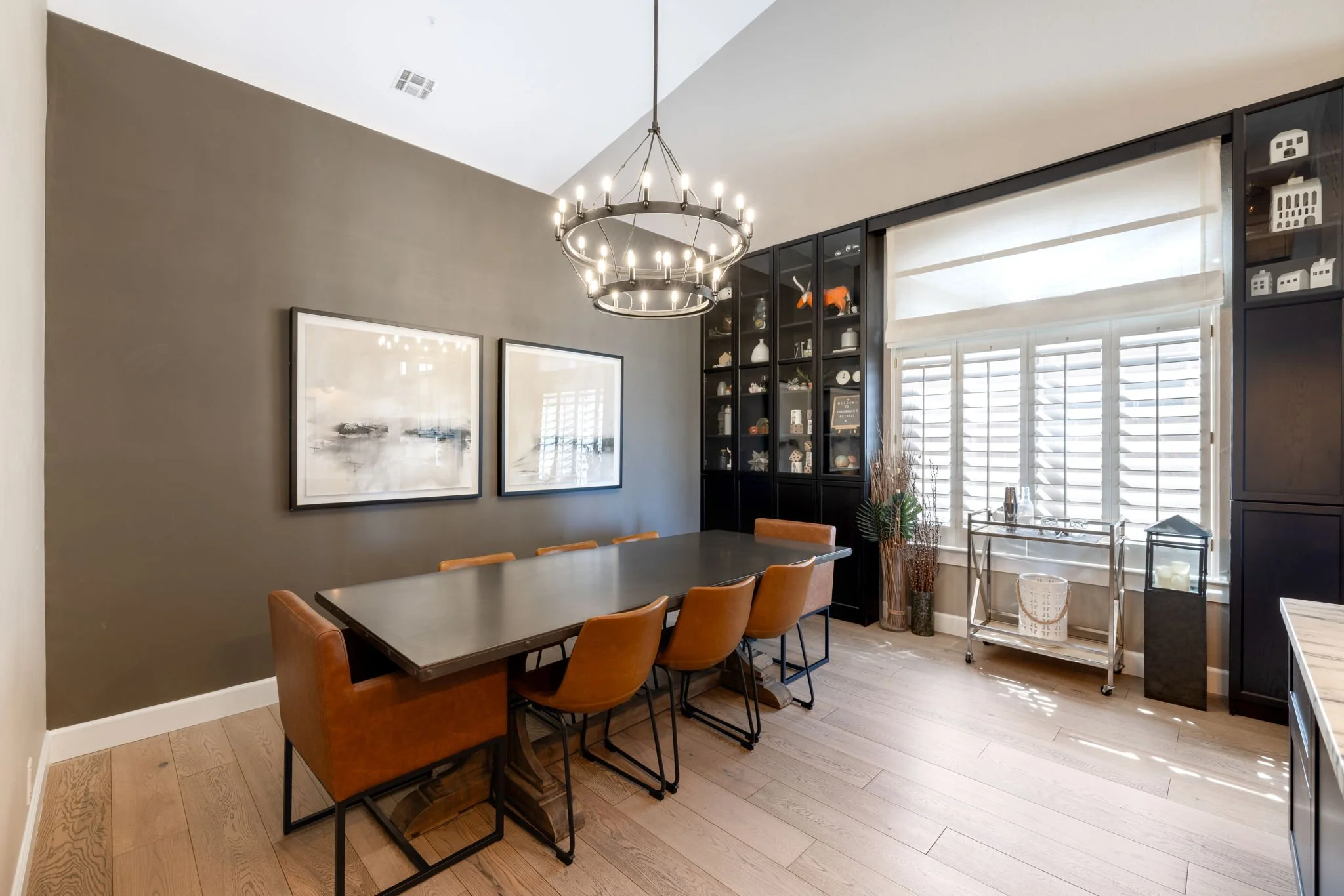Modern dining room with a black table, six brown leather chairs, a black shelving unit filled with decorative items, and a large window with white shutters and blinds, all illuminated by a circular chandelier.