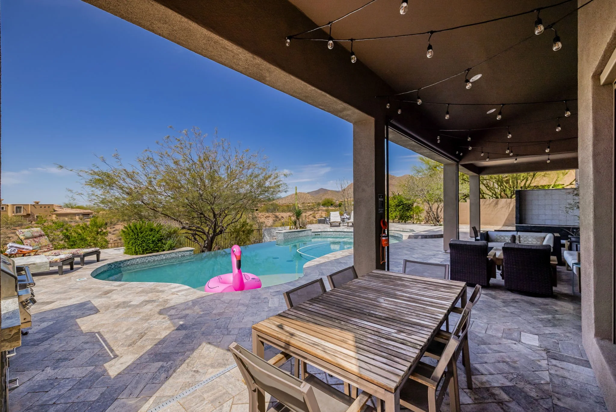 Outdoor patio with a swimming pool, inflatable pink flamingo, and desert landscape with mountains in the background.