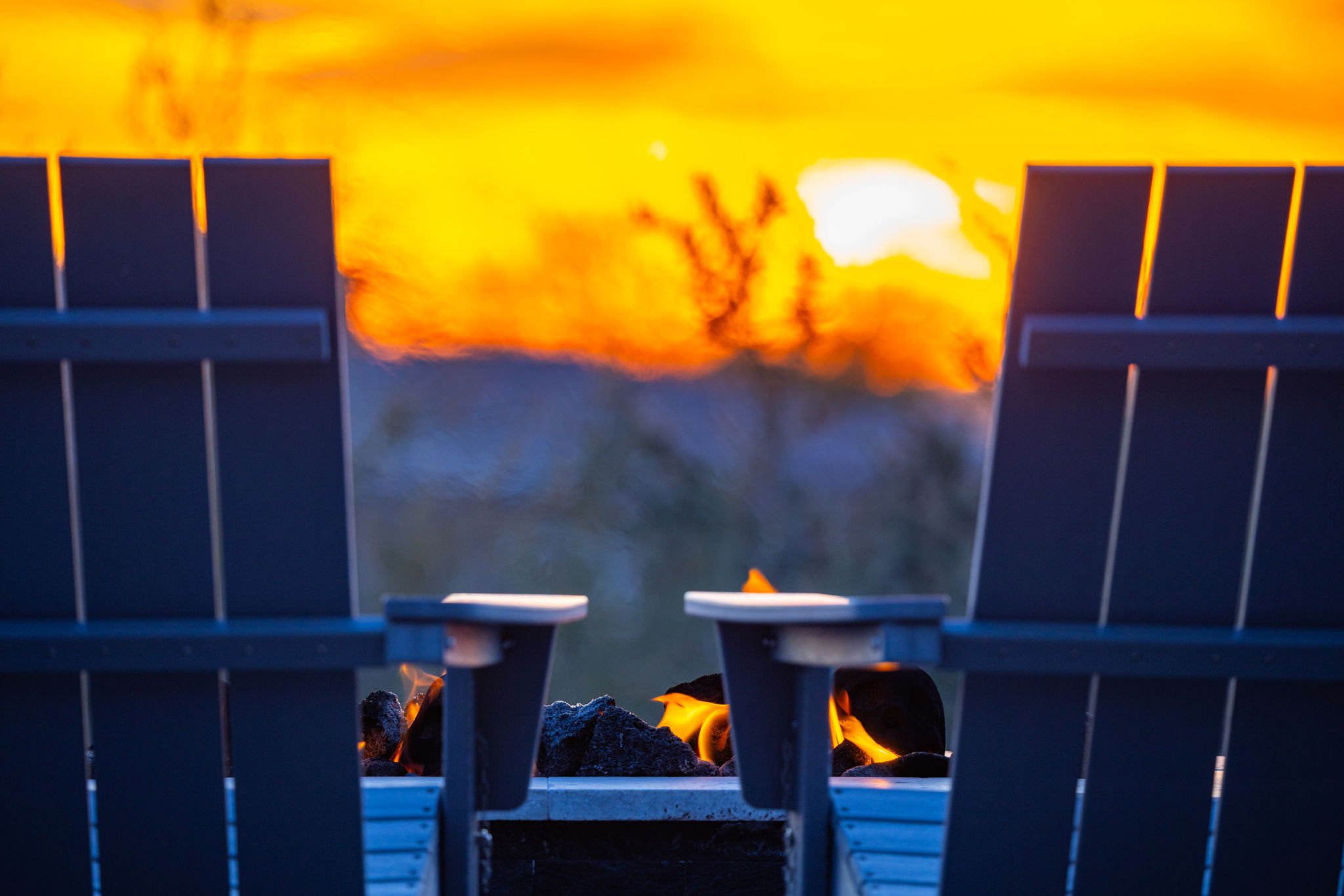 Two Adirondack chairs facing a sunset with the sky glowing orange and yellow. A firepit with visible flames sits between the chairs.