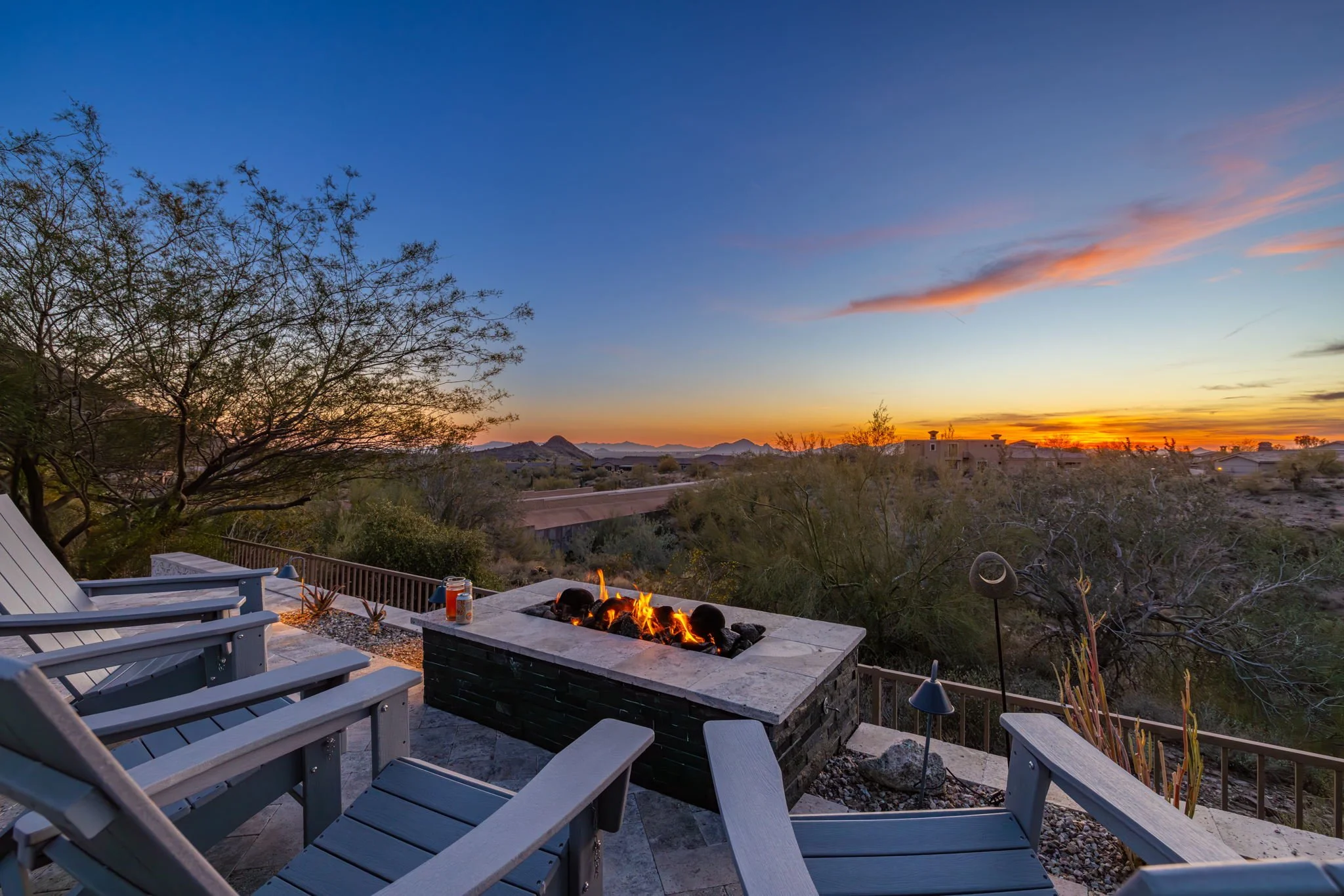 An outdoor patio area with a stone fire pit and chairs, overlooking a desert landscape at sunset.