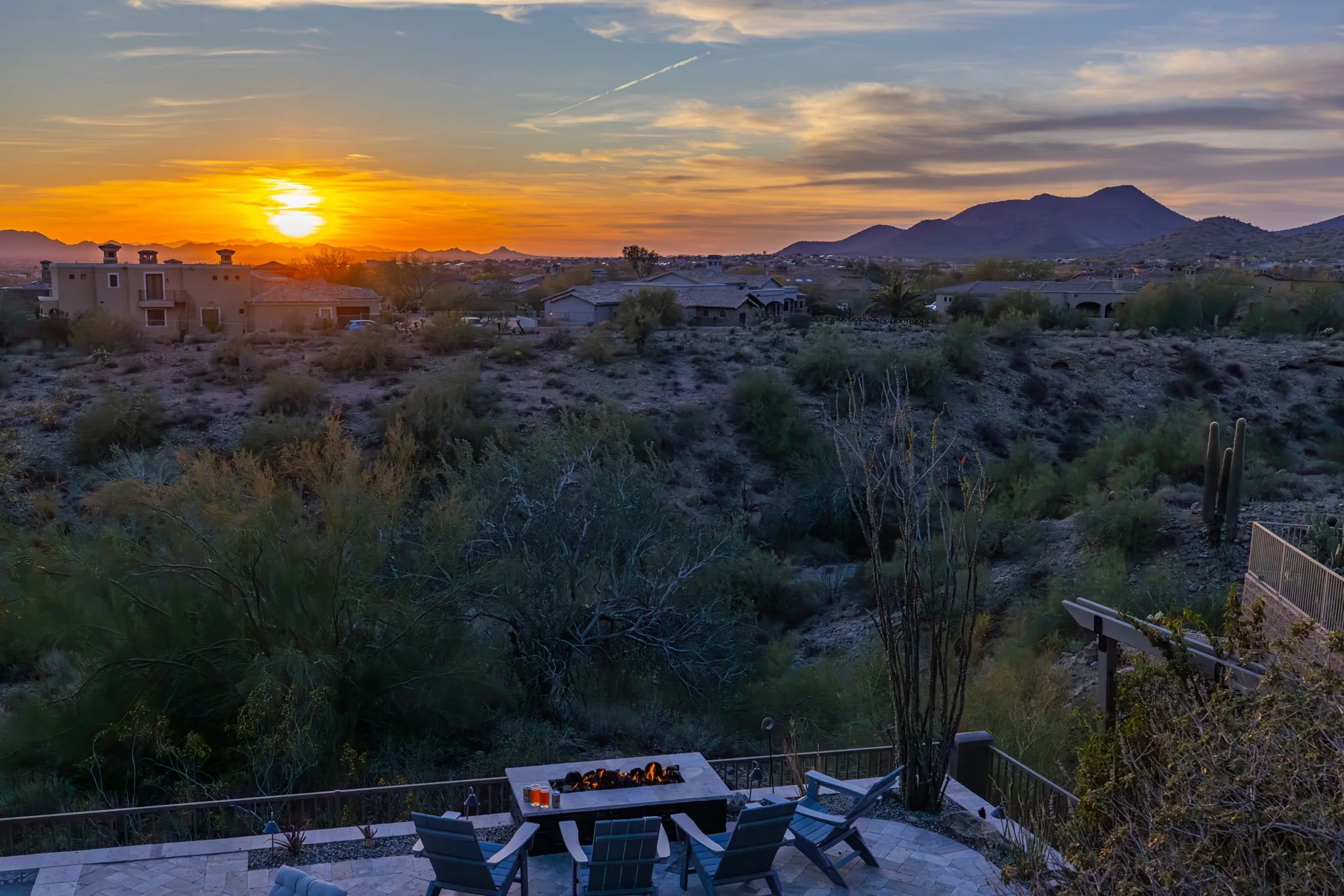 Sunset over a desert landscape with mountains in the distance and houses with tiled roofs. In the foreground, a patio with chairs and a fire pit.