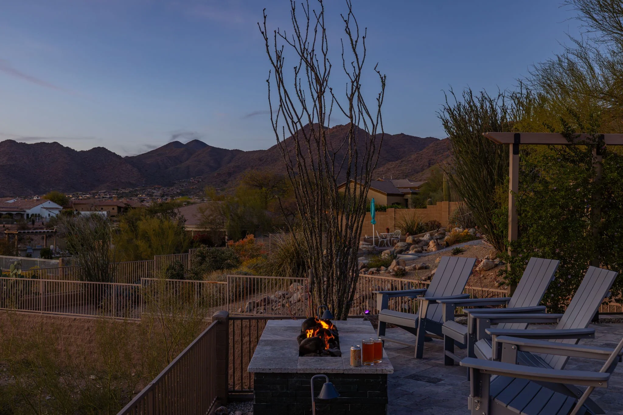 A backyard patio with a fire pit, three chairs, some drinks, and a view of the mountains at dusk.