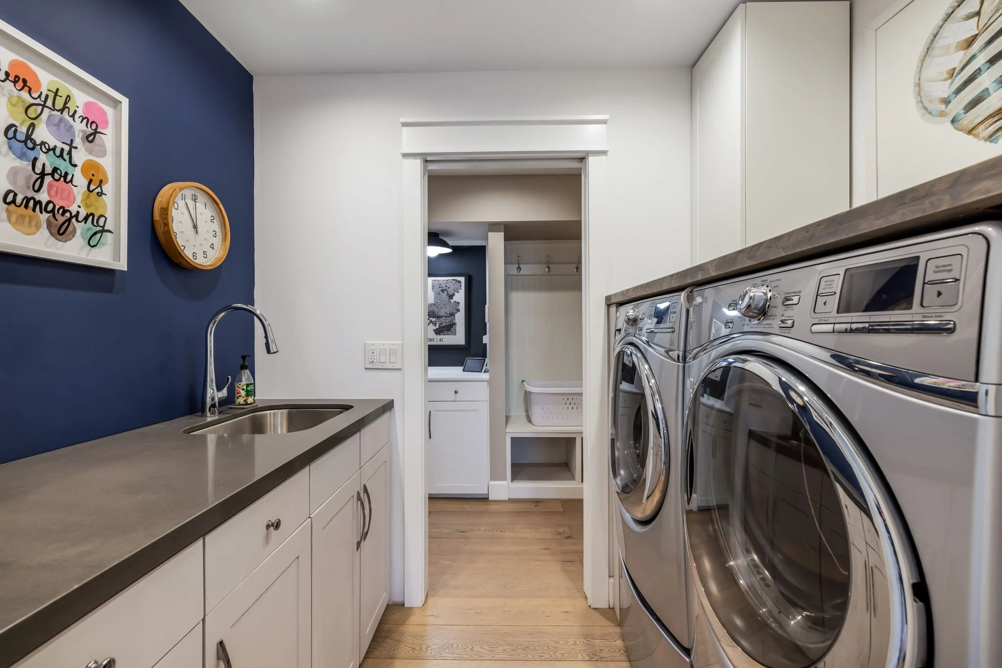 A laundry room with a white cabinet, a gray countertop, a stainless steel sink with a faucet, and a blue accent wall featuring a colorful wall art and a round clock. On the right, there are stacked front-loading washing and drying machines, and there