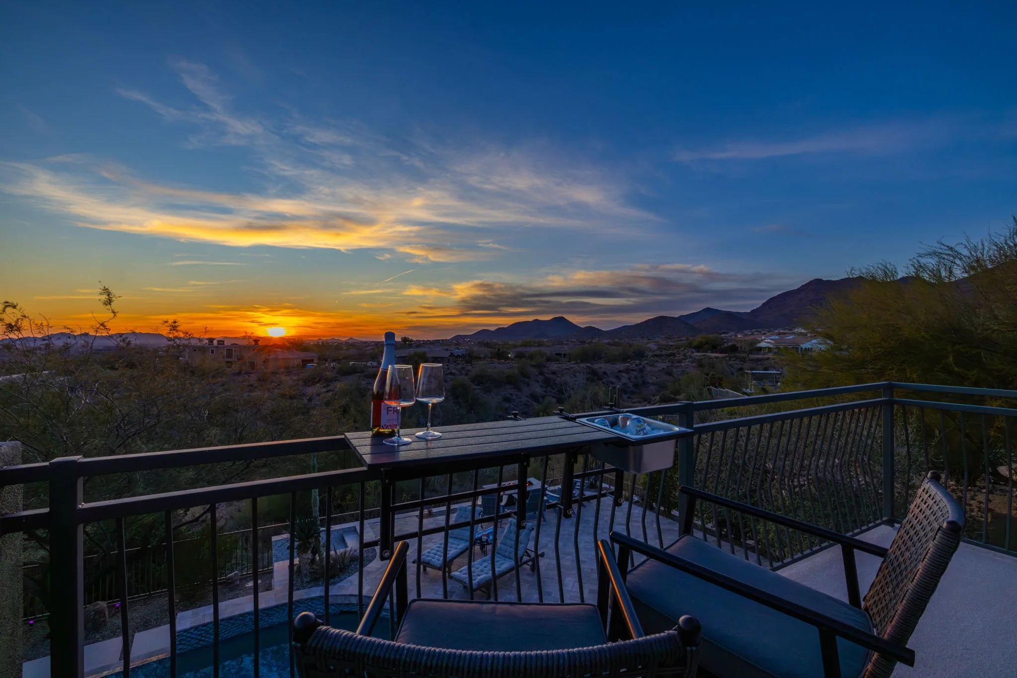 Balcony overlooking desert landscape at sunset with a table, wine bottle, and two glasses.
