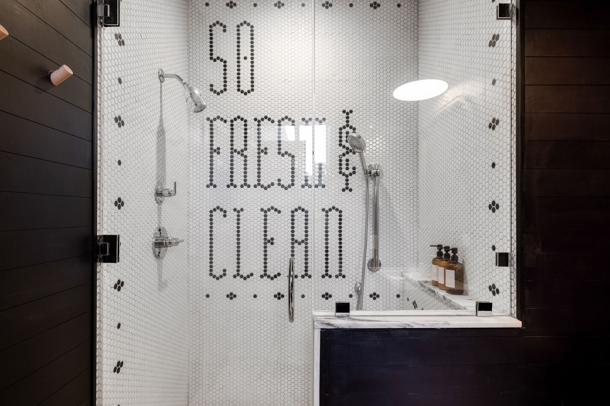 Shower area with white hexagonal tiles, black dot accents, and the words 'Stay Fresh Clean' spelled out in black tiles on the wall. Contains ceiling-mounted showerhead, adjustable handheld shower, and three amber soap bottles on a ledge.