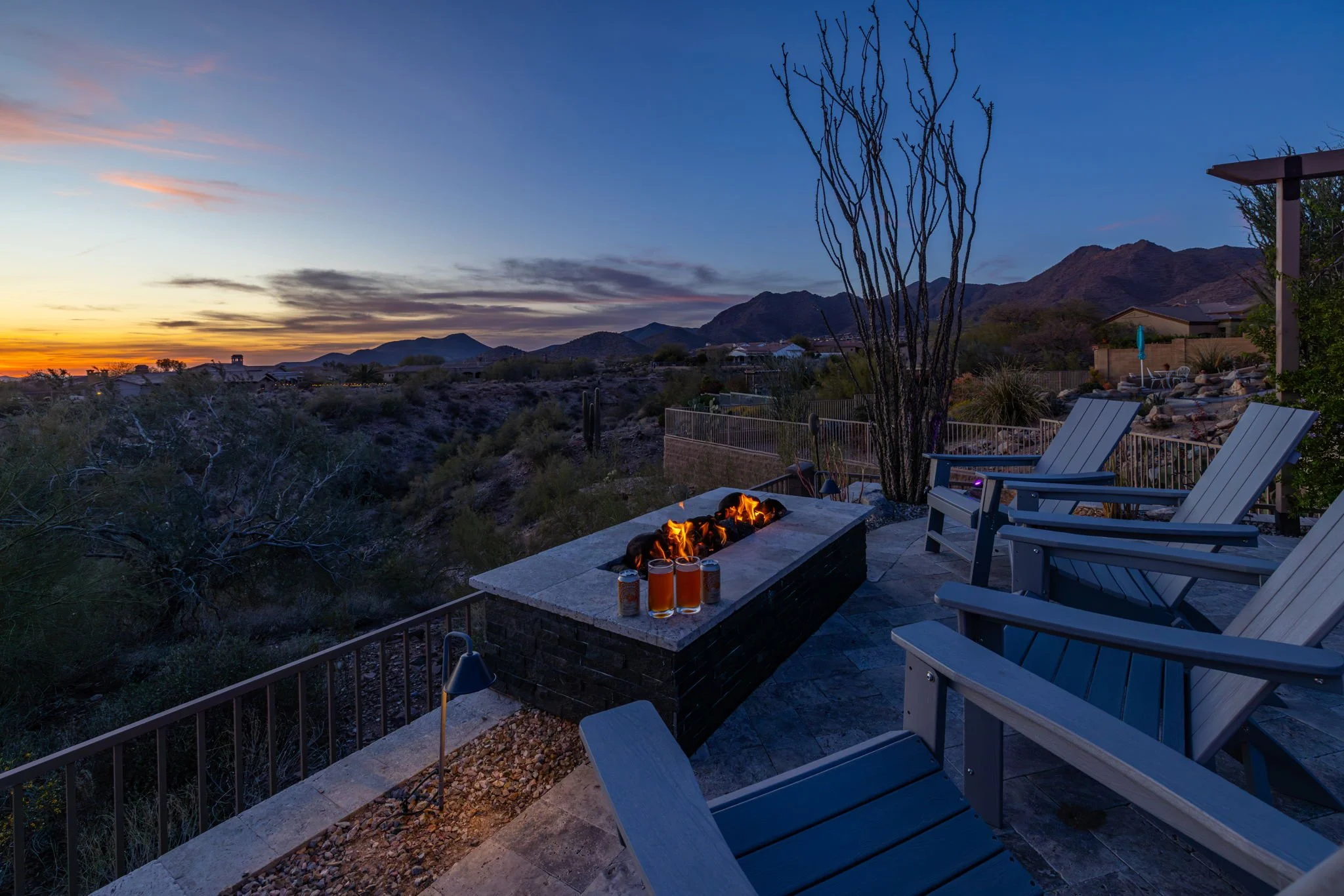 A backyard patio during sunset with a fire pit, drinks, Adirondack chairs, and desert mountains in the background.