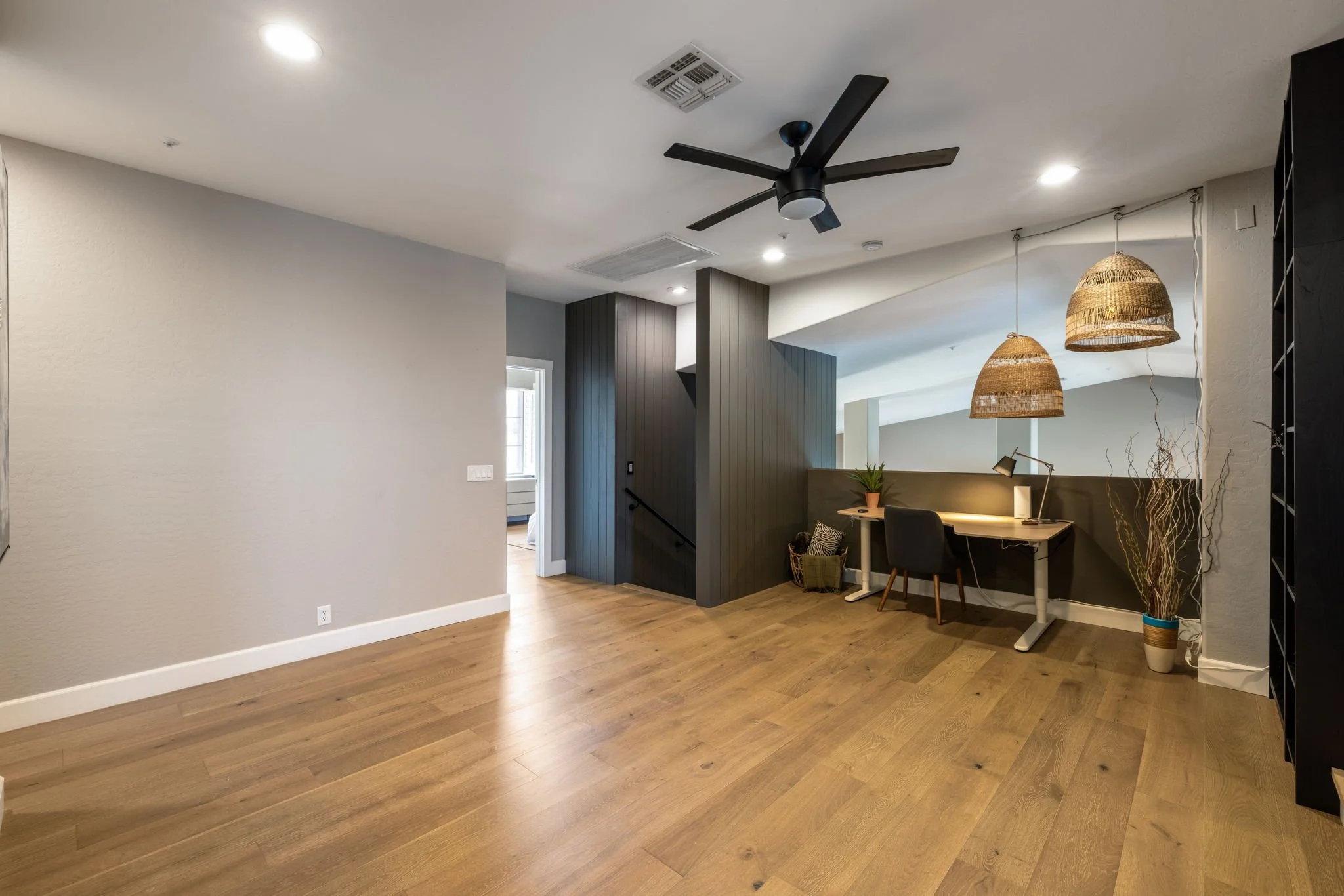 Empty living space with wood flooring, light gray walls, a black ceiling fan, a small workspace with a desk, chair, and two hanging woven lamps, and a door leading to another room.