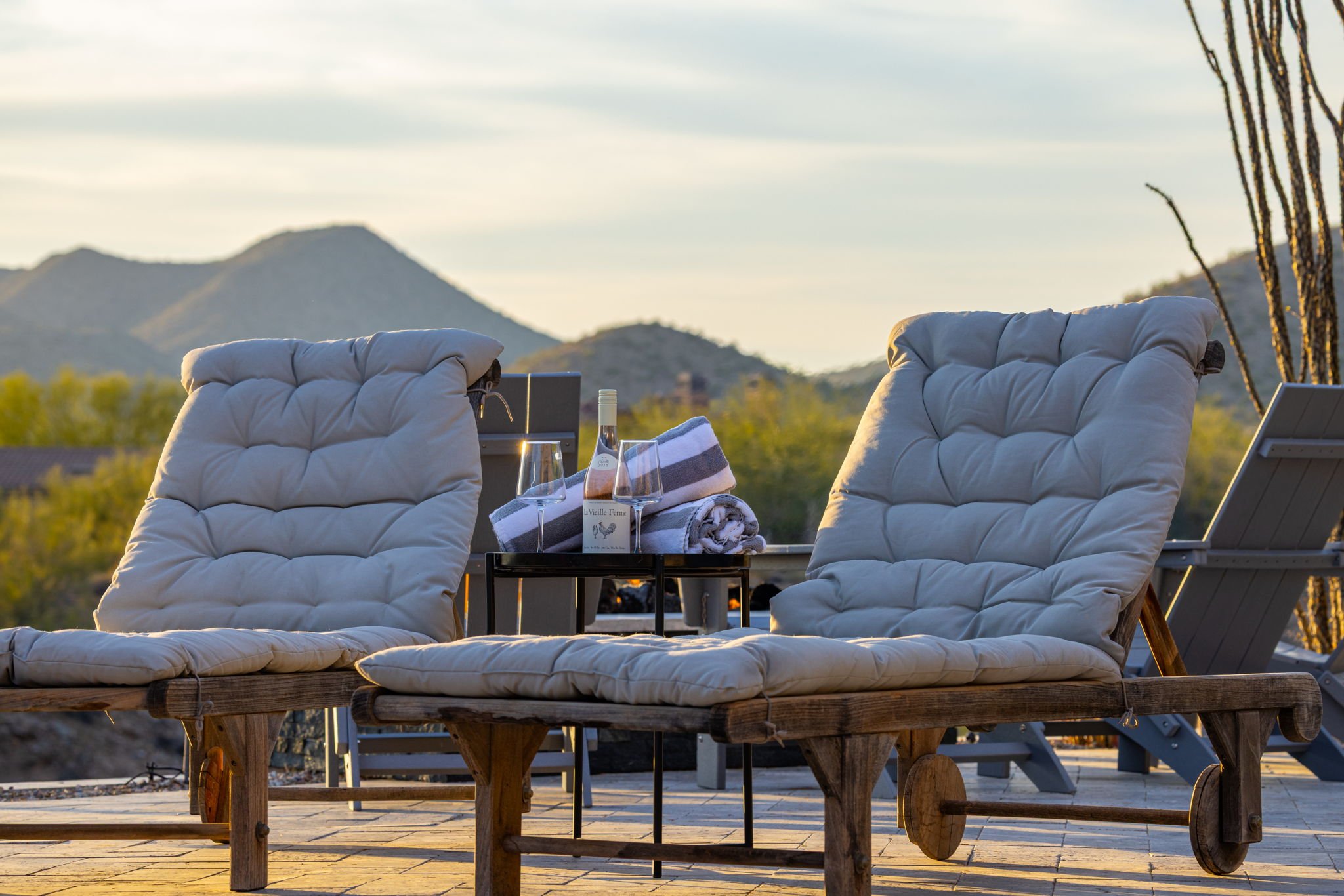 Outdoor patio seating with beige cushioned lounge chairs, a small black table with a bottle of rosé, two wine glasses, towels, and a desert mountain landscape in the background at sunset.