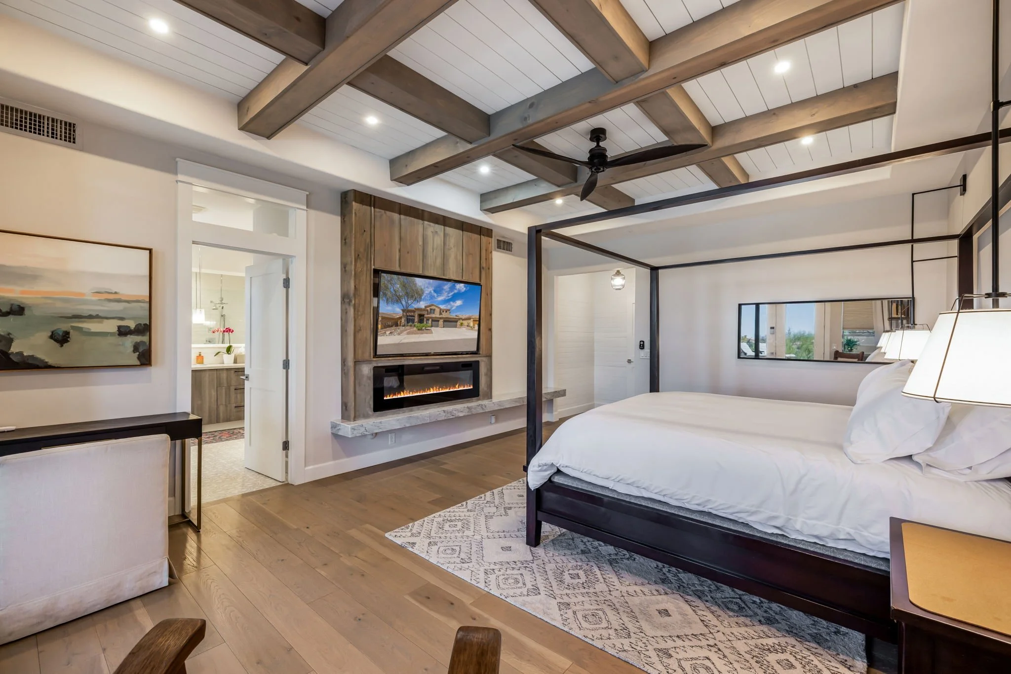 A modern bedroom with a white canopy bed, wooden beams on the ceiling, a wall-mounted television above a fireplace, and a window with a view outside.