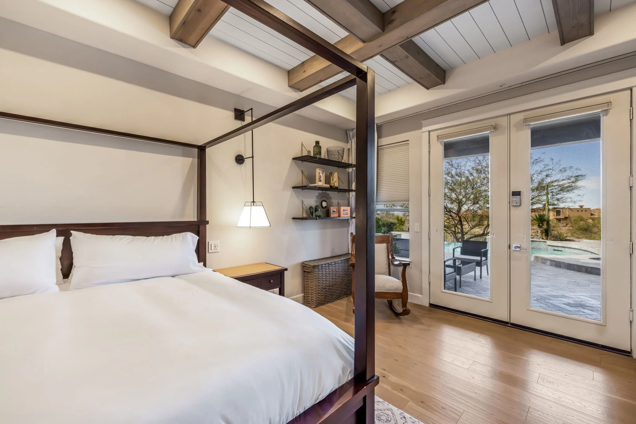 Bedroom with a four-poster bed, bedside table, hanging lamp, shelves with decorative items, and a glass door leading to an outdoor patio with outdoor furniture and a pool, overlooking trees and a desert landscape.