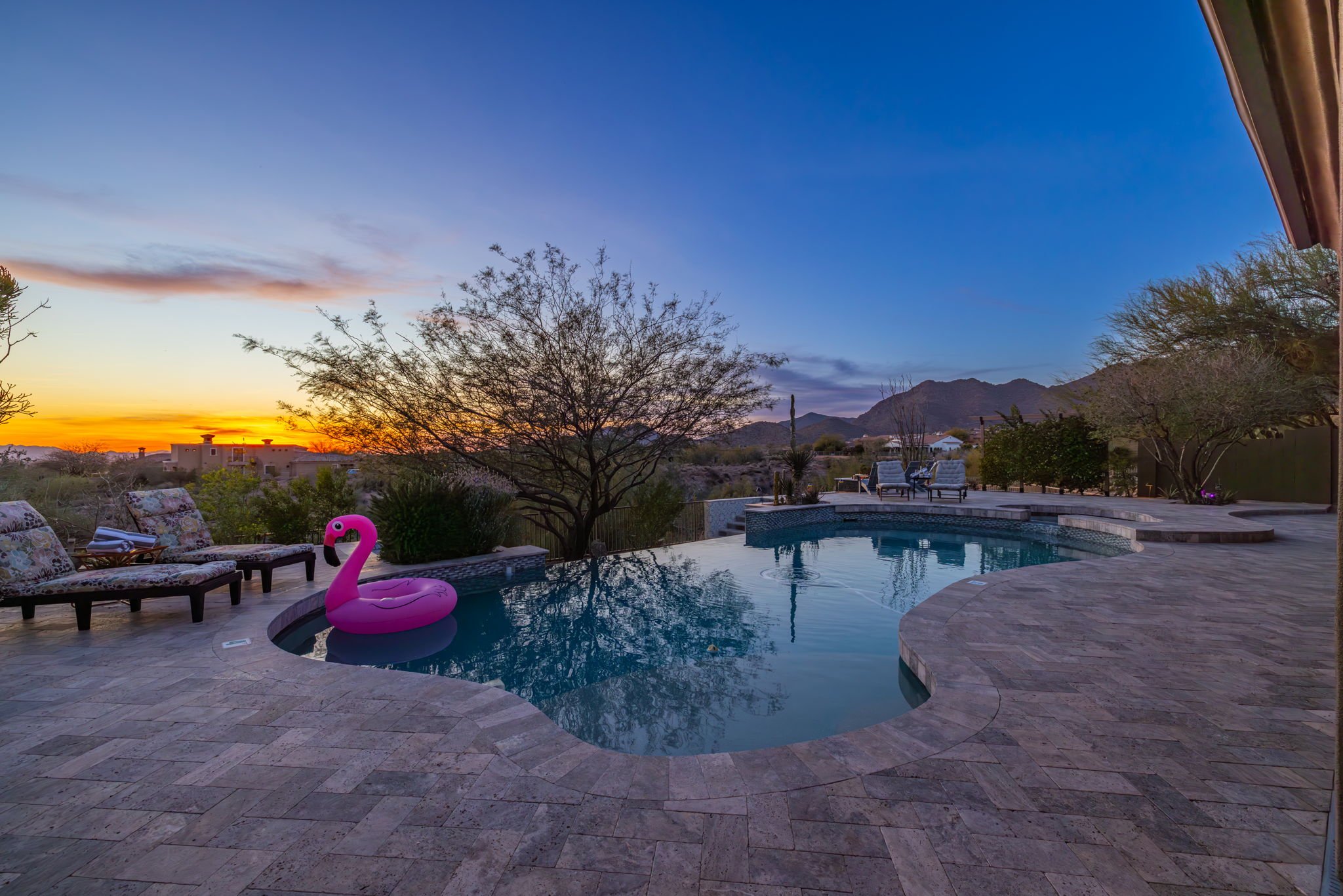 A backyard swimming pool area at sunset with mountain views, a pink flamingo float in the pool, and outdoor seating including chairs and lounge chairs.