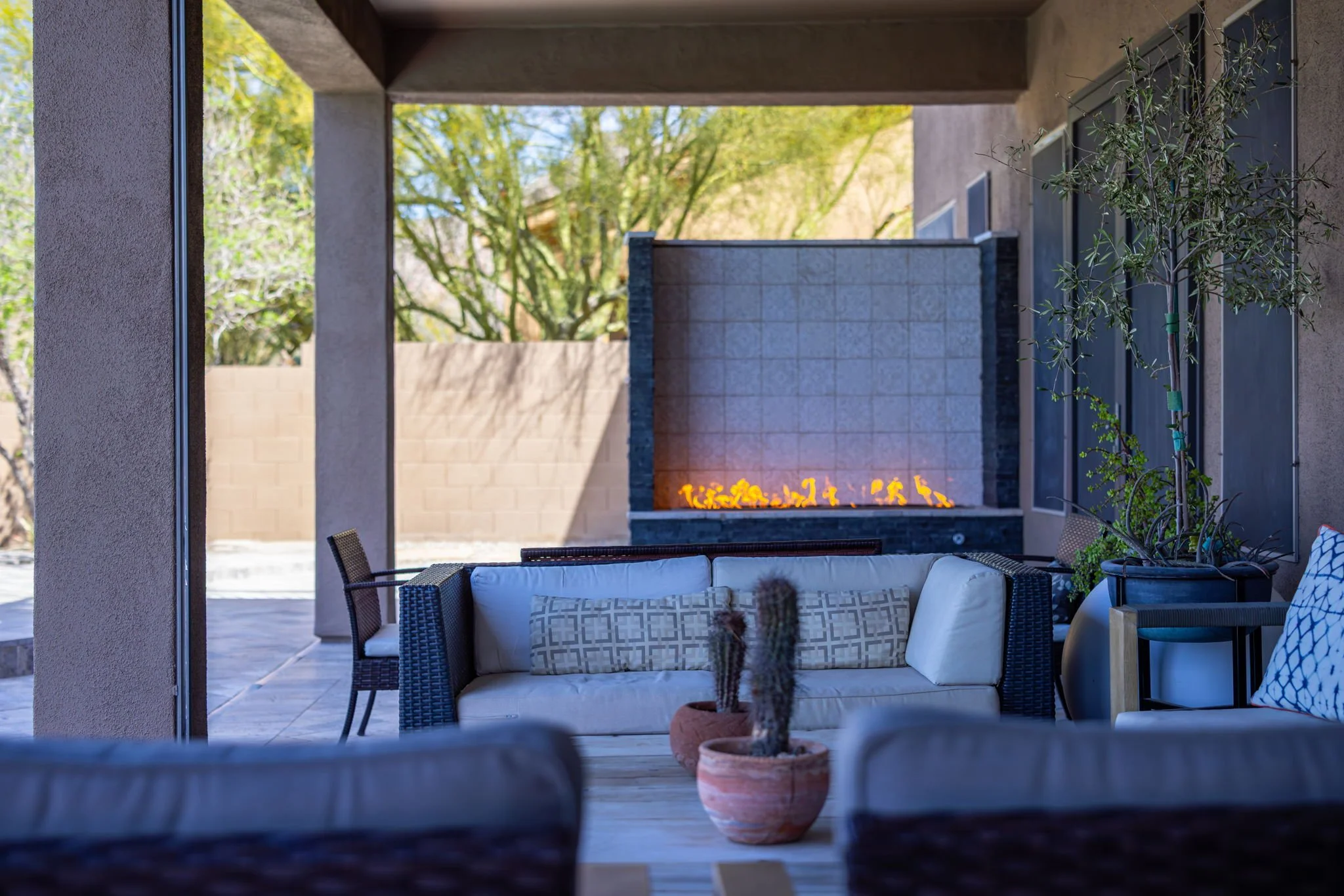 Outdoor patio with a comfortable white sofa, potted cacti, and a modern water feature with fire, under a covered patio with trees in the background.