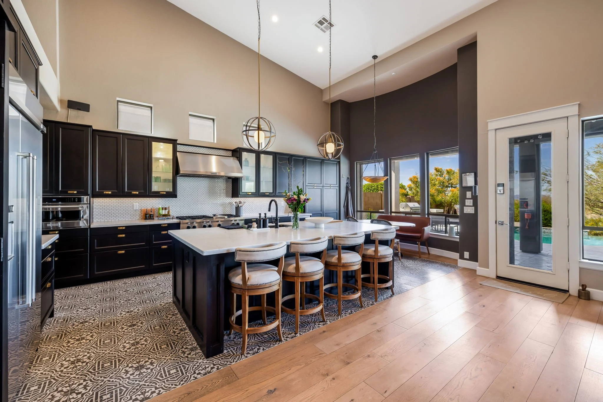 Modern kitchen with black cabinets, white countertops, patterned tile flooring, and large windows showing outdoor greenery.