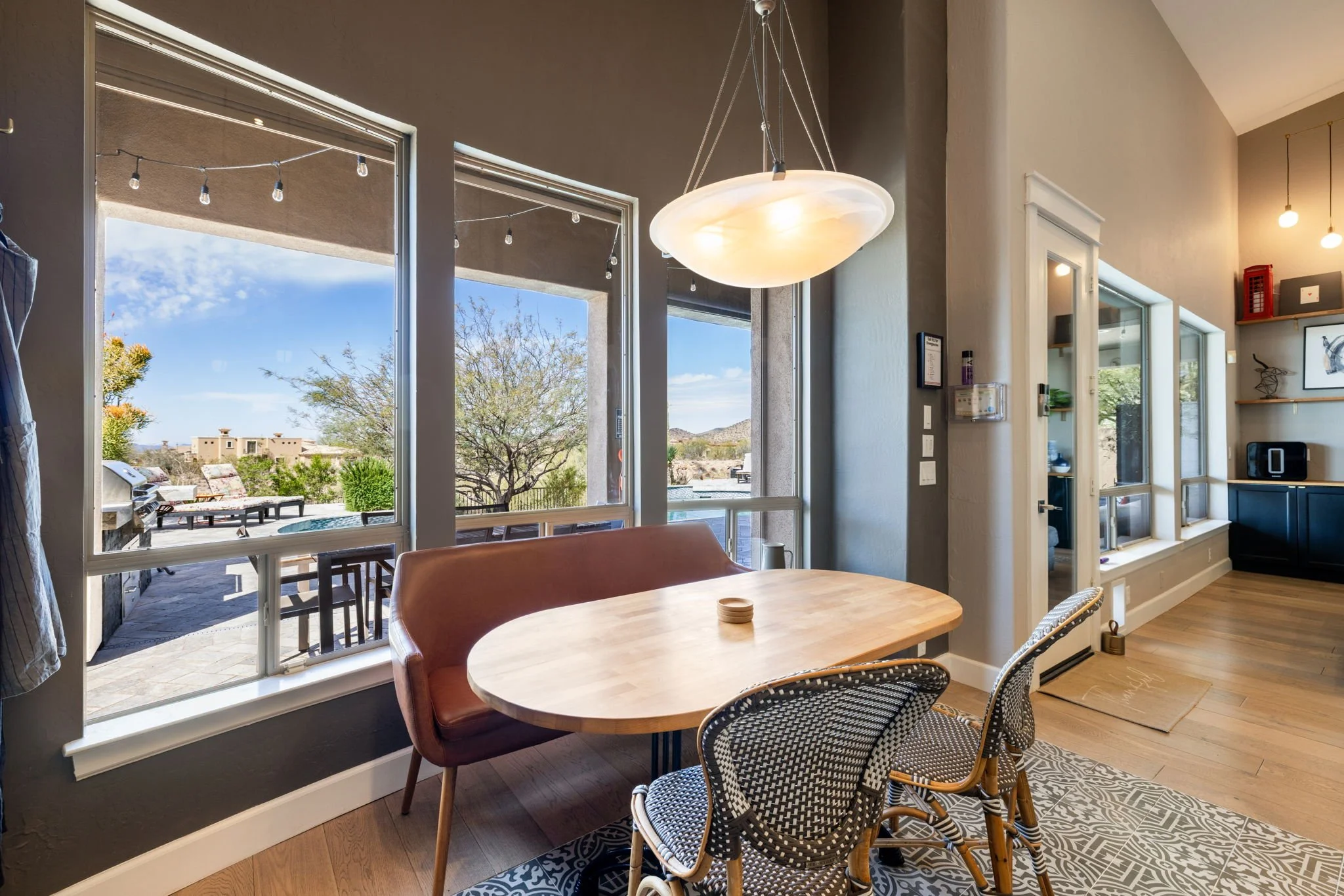 Dining area with a wooden table, two patterned chairs, a leather bench, large windows showing a patio with outdoor furniture, trees, and a blue sky, and a modern hanging light fixture.