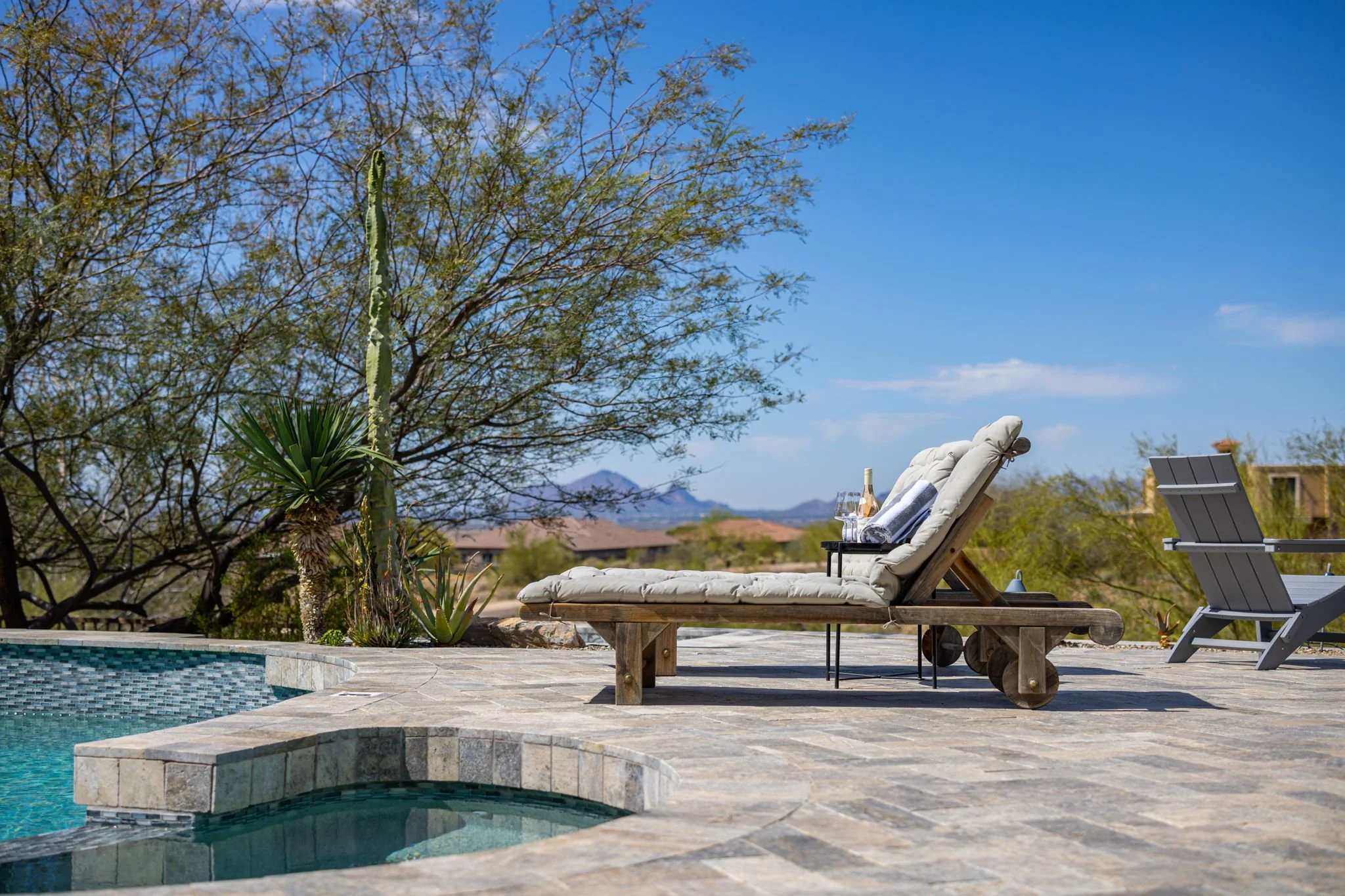 Poolside lounge chair with cushions, drinks, and towels on a stone deck, with desert plants and trees in a sunny outdoor setting.