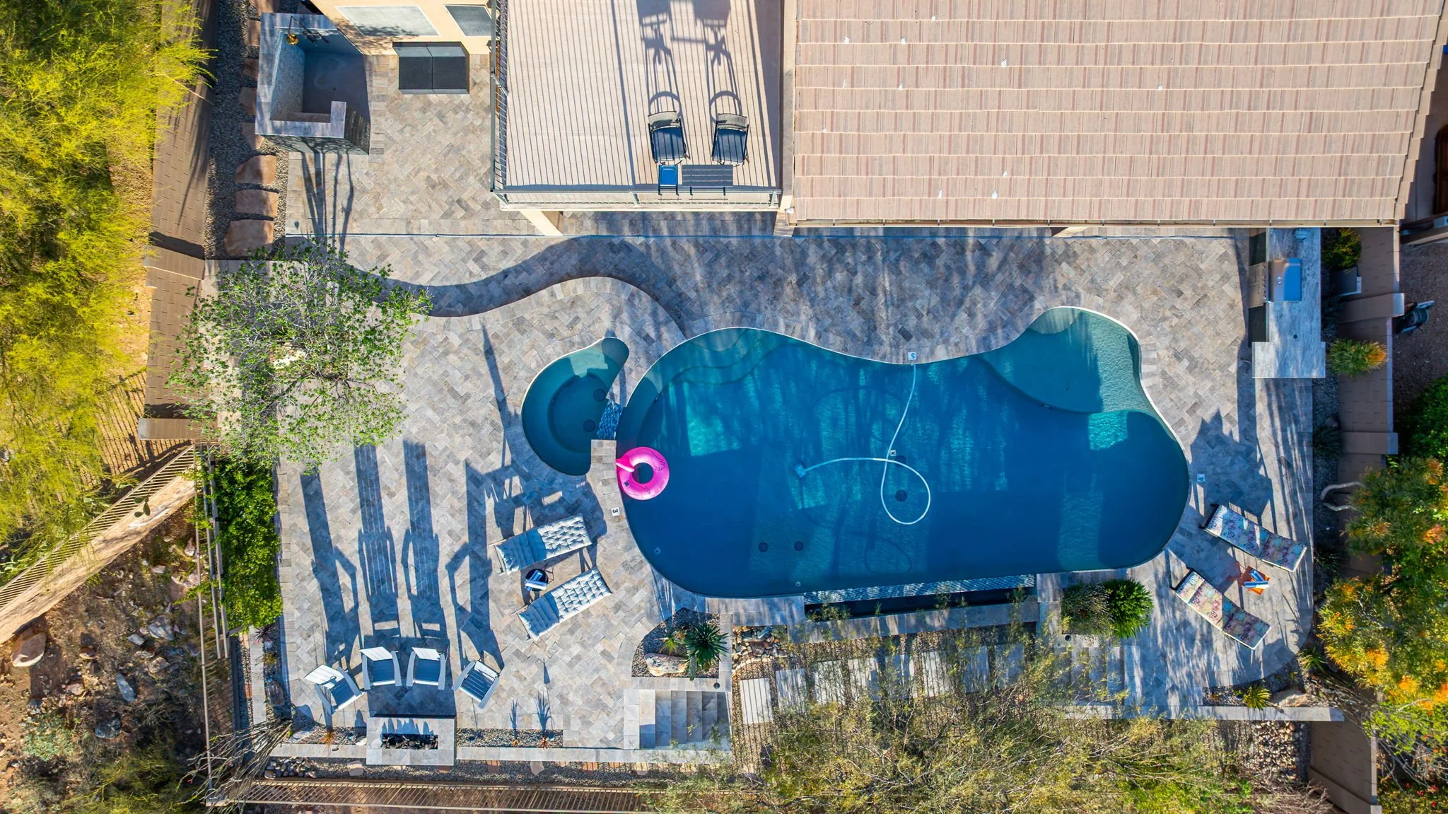 Aerial view of a backyard with a freeform swimming pool, lounge chairs, a shaded seating area, and surrounding greenery.