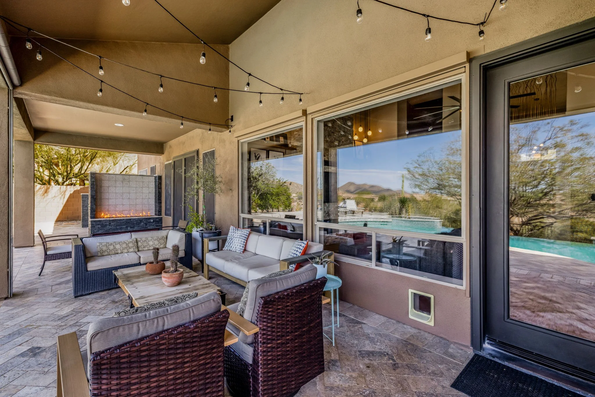 Covered outdoor patio with string lights, outdoor fireplace, patio furniture including sofas and chairs with cushions, a side table, large windows, a sliding glass door, and a view of a swimming pool and desert landscape with mountains in the backgro