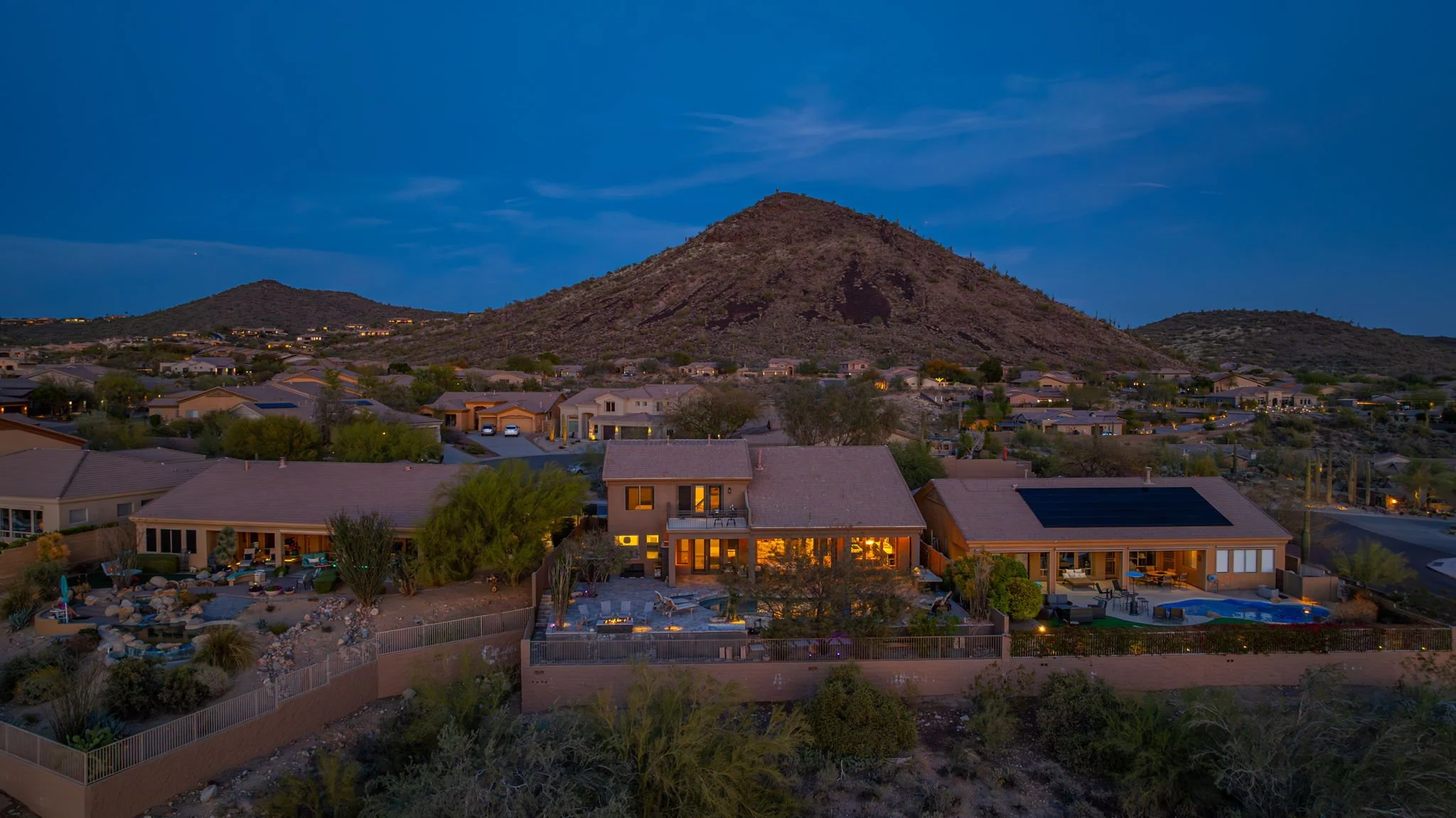 Night view of a suburban neighborhood in a desert landscape, with a mountain in the background. Houses are illuminated with interior and exterior lights, some featuring swimming pools and outdoor seating areas.