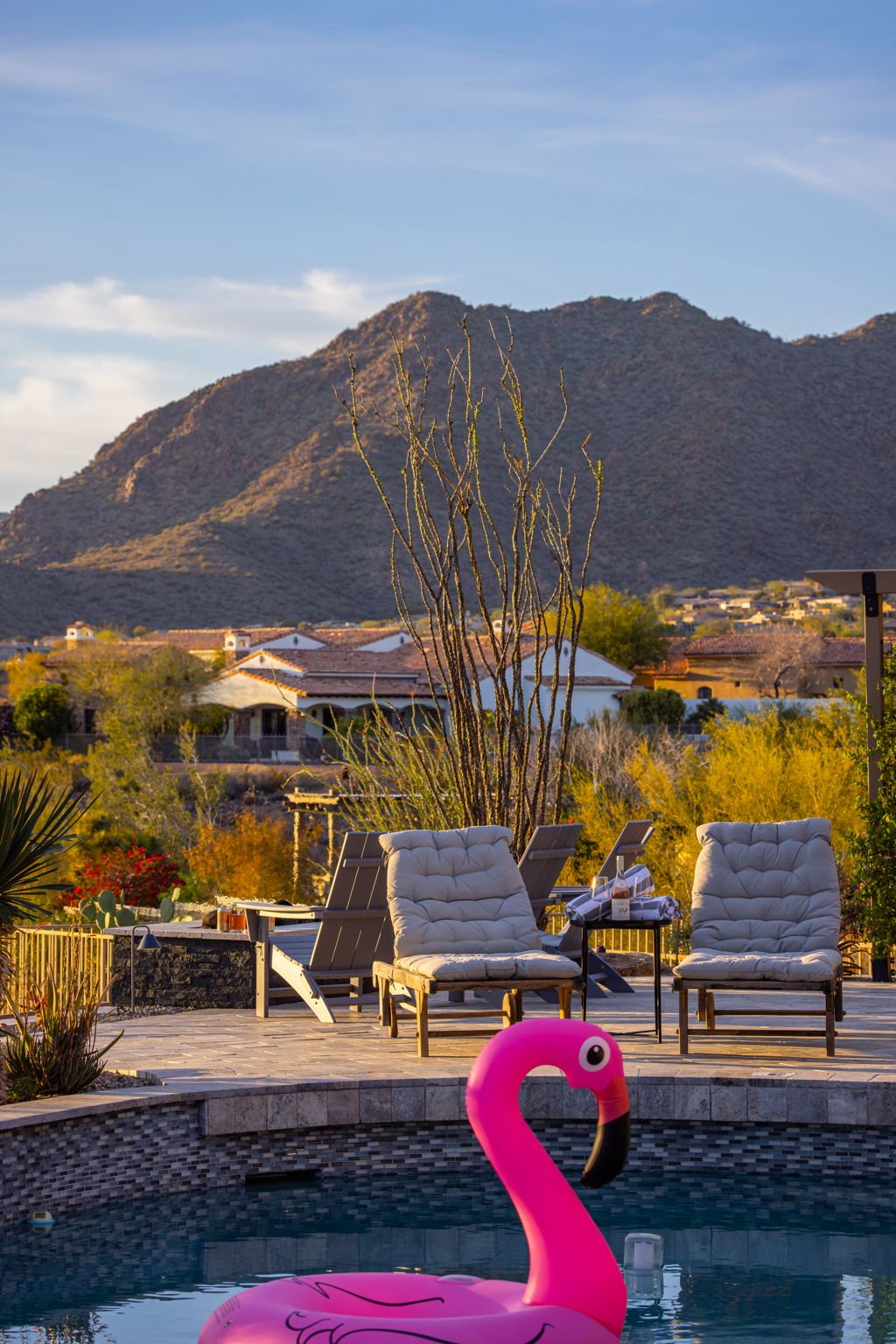 A backyard swimming pool with a pink flamingo float, outdoor chairs, and a desert mountain landscape in the background during sunset.