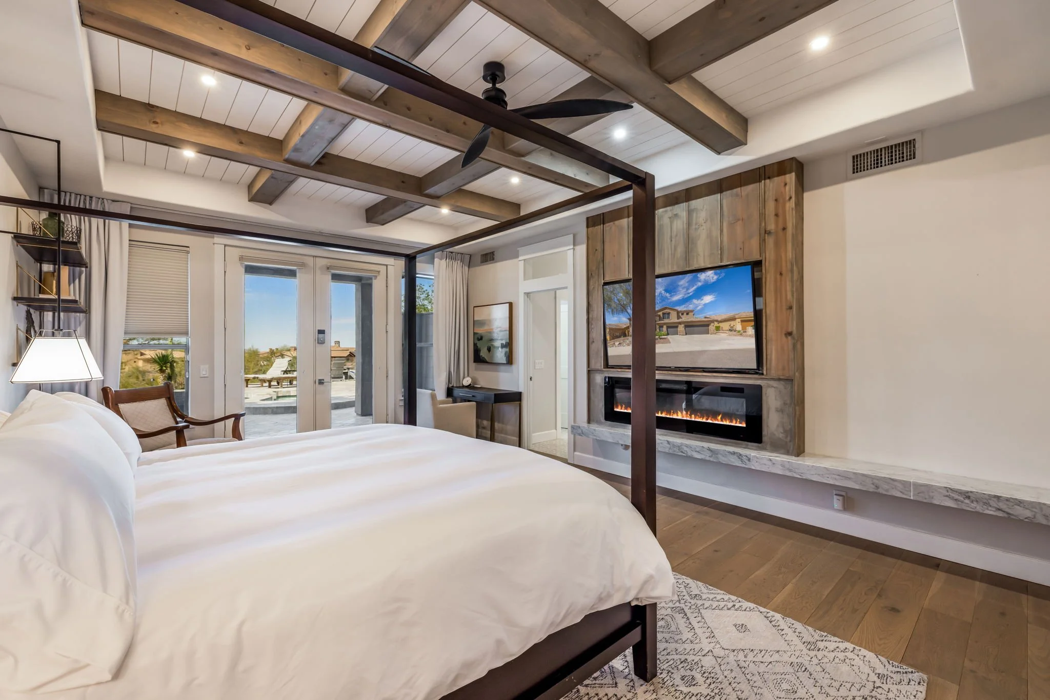 Modern bedroom with a four-poster bed, a wall-mounted TV above a fireplace, a ceiling fan, and large glass doors leading to a balcony with outdoor seating.