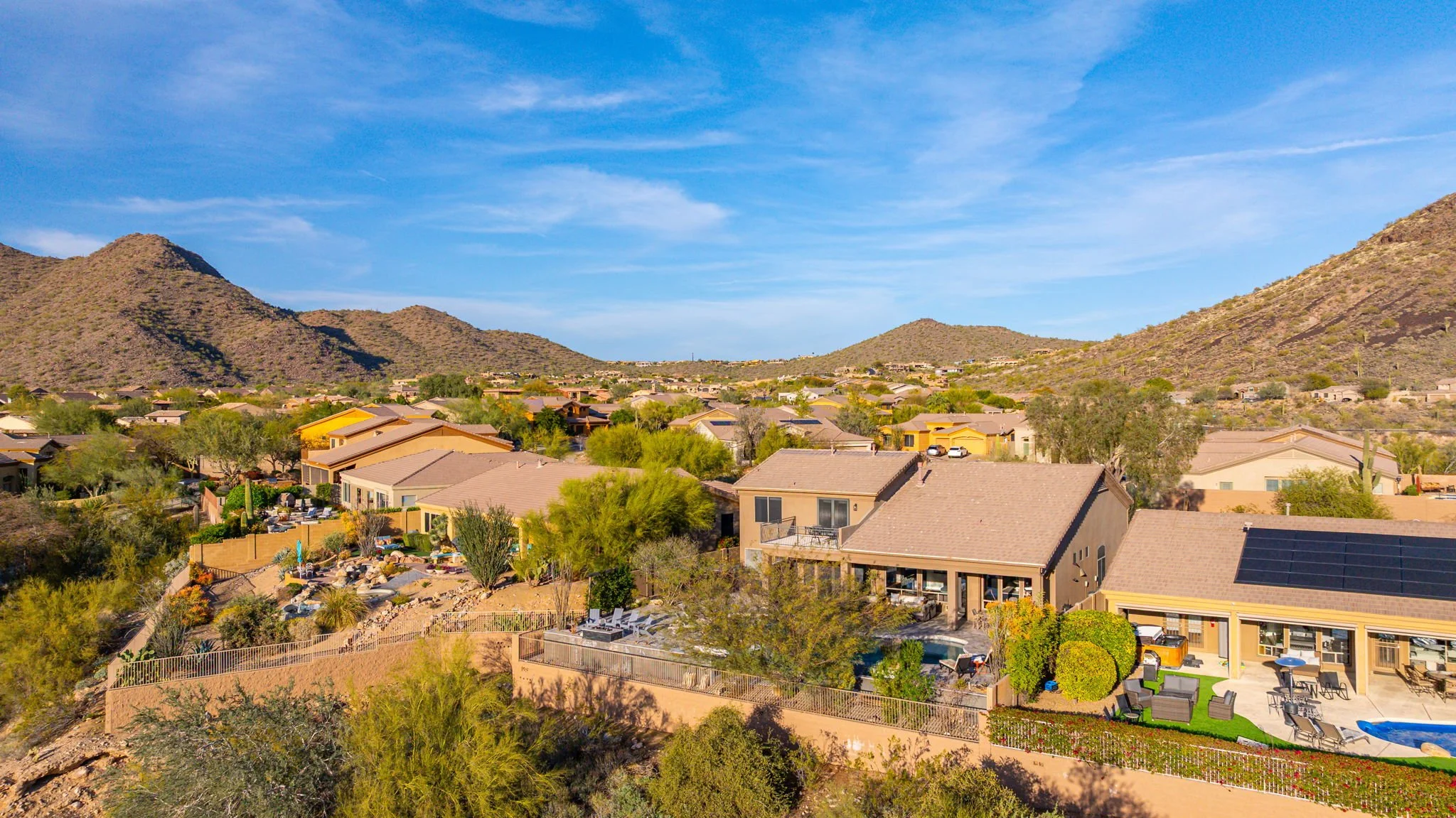 Aerial view of a suburban neighborhood in a desert landscape with mountains in the background, featuring houses with tan and yellow exteriors, some with solar panels, and backyards with outdoor furniture, trees, and landscaping.