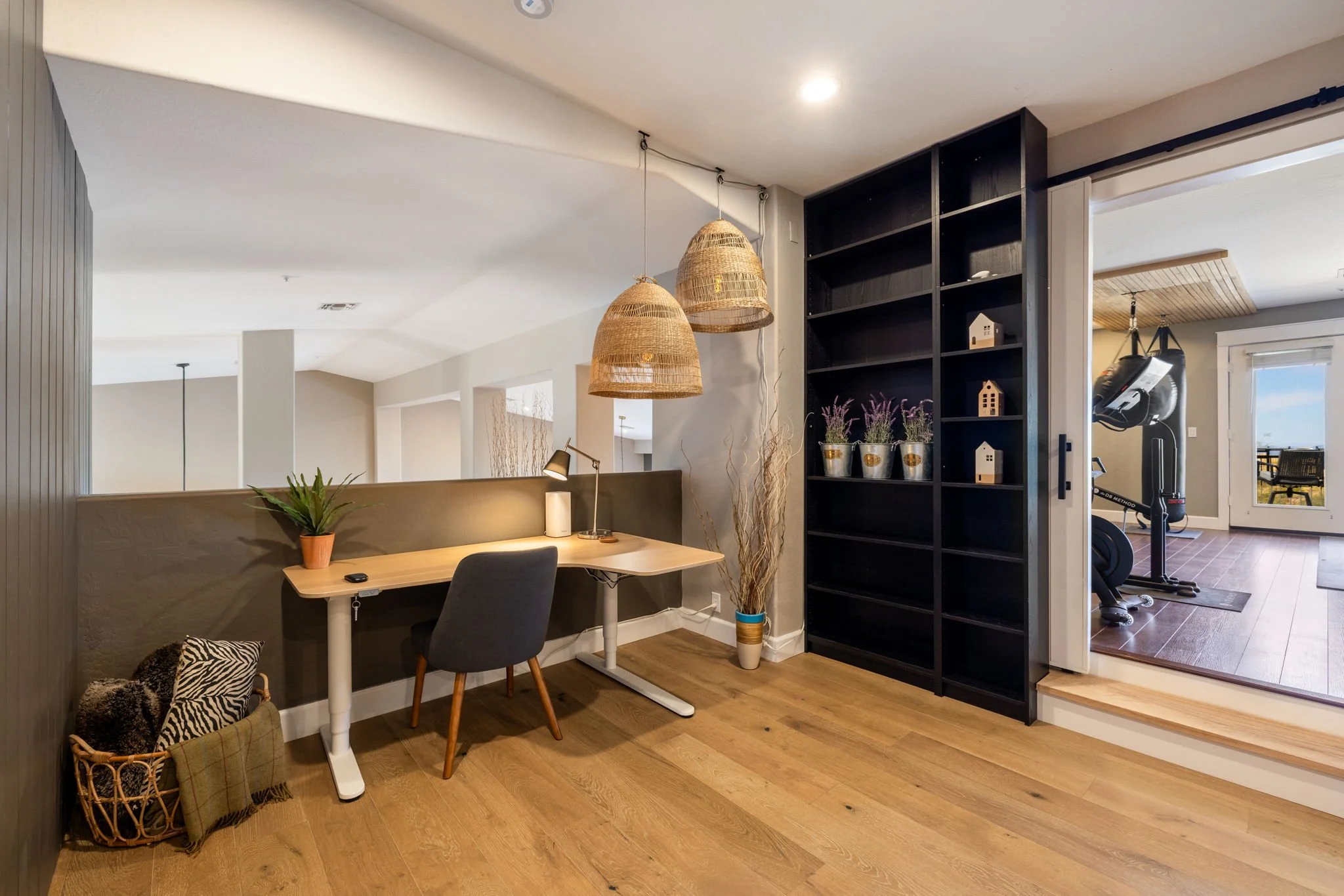 Home office with a desk, black office chair, potted plants on a shelf, hanging wicker lamps, and a basket of throw blankets. Adjacent is a fitness area with a punching bag and exercise equipment, separated by a sliding door.