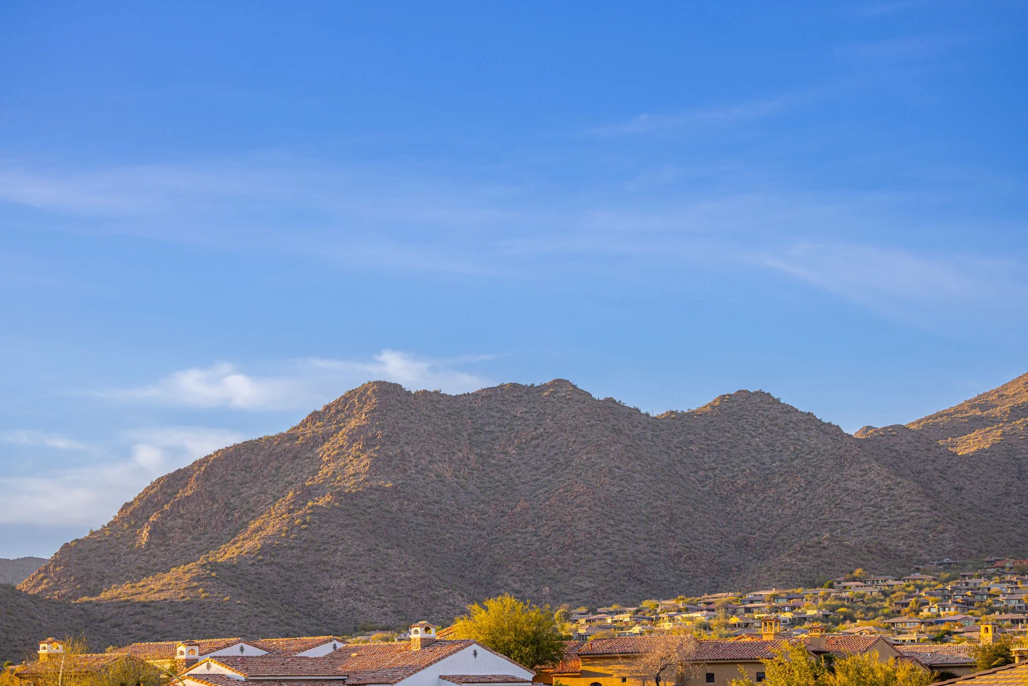 A scenic view of mountains with a small town in the foreground, featuring terracotta roofs and trees, under a blue sky with a few clouds.