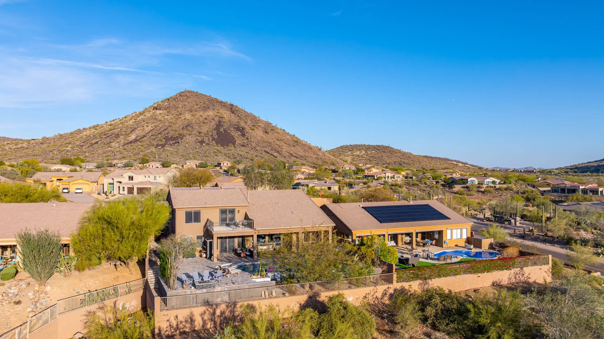 A residential neighborhood with houses, some equipped with solar panels, set against desert mountains under a clear blue sky.