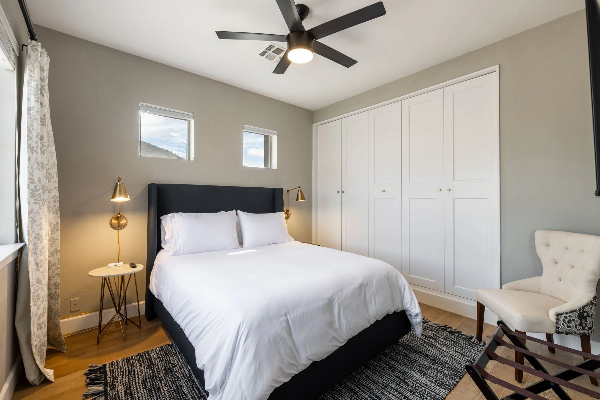 A modern bedroom with a white bed, black headboard, gold wall lamps, a small side table, white closet doors, a beige upholstered chair, and part of a window with patterned curtains.