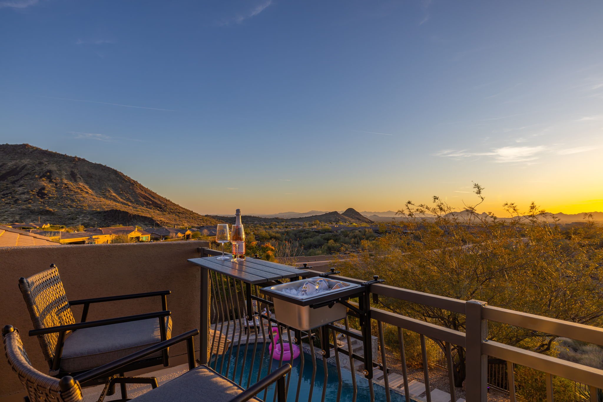 A balcony with wicker chairs and a table holding a bottle of wine and two wine glasses, overlooking a desert landscape with hills at sunset.