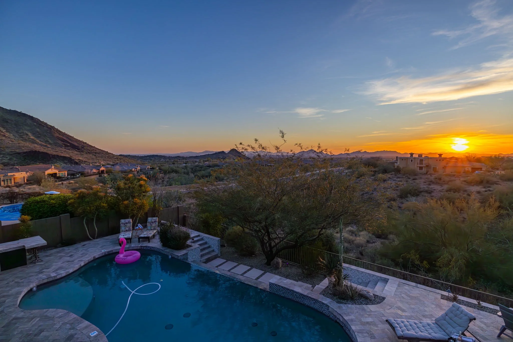 A backyard swimming pool with a pink flamingo float, surrounded by patio furniture and trees, overlooking a desert landscape at sunset with mountains in the background.