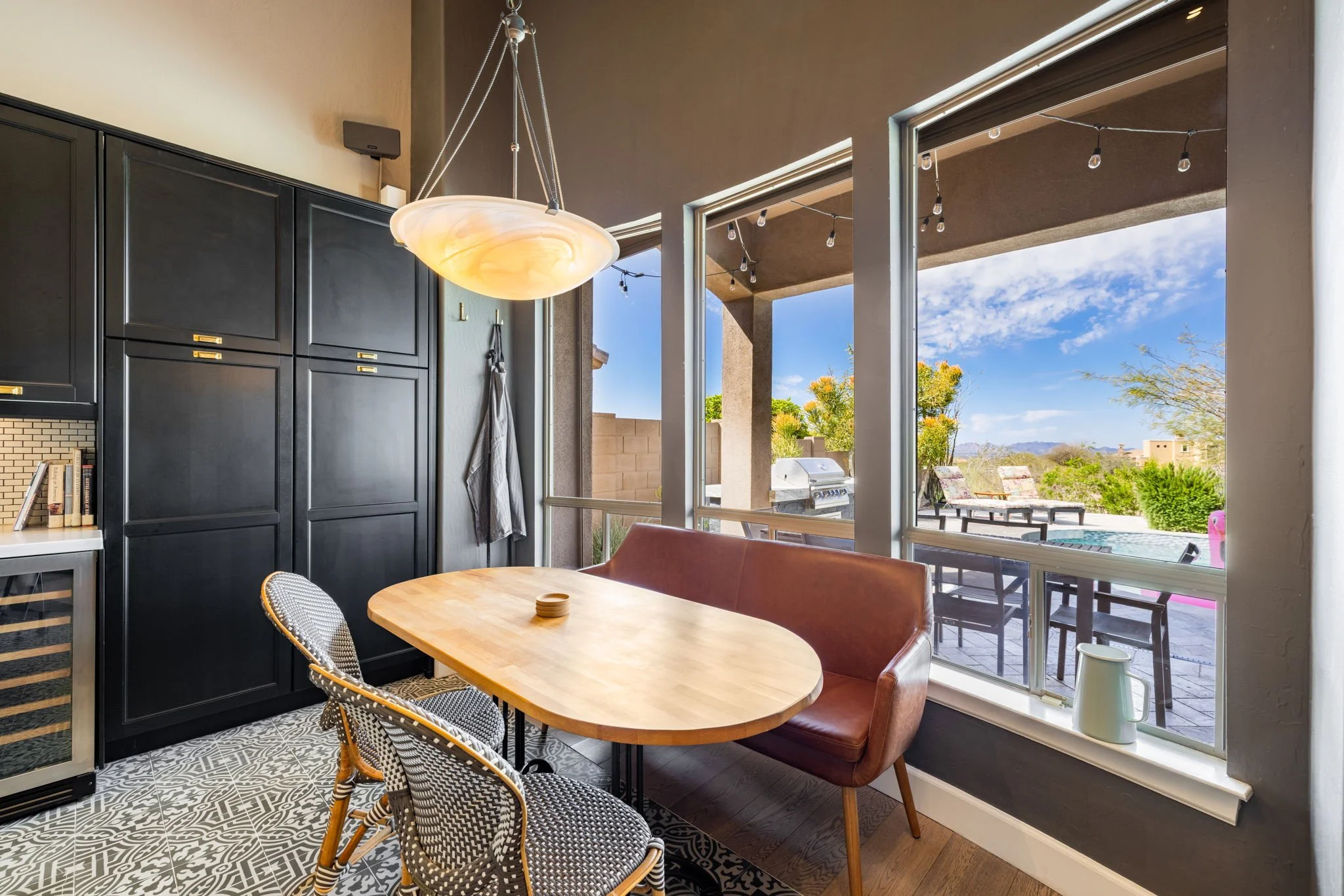 Kitchen with black cabinets, wooden table, patterned chairs, and large windows overlooking a patio with outdoor seating and greenery under a blue sky.