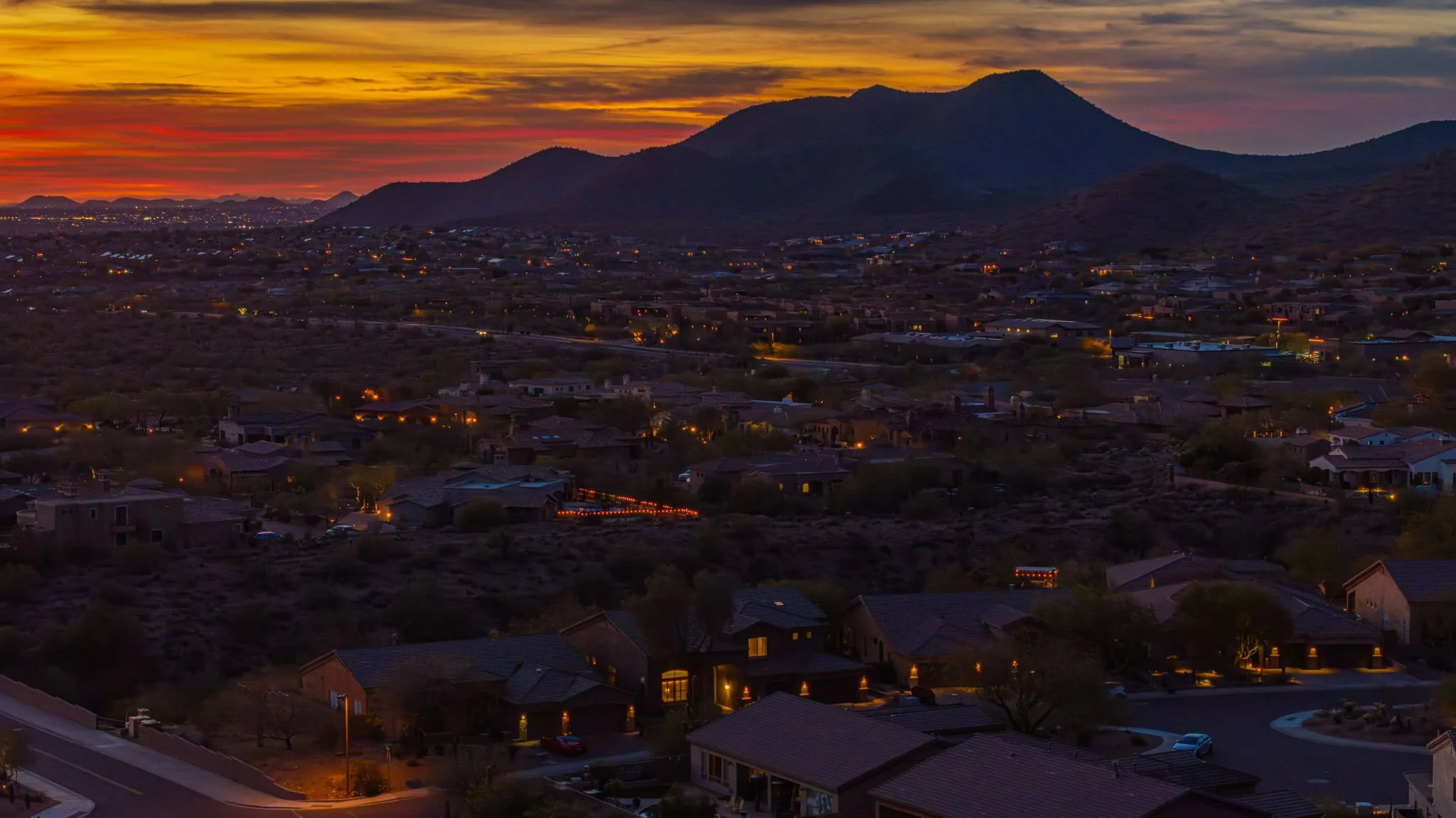 A sunset over a suburban neighborhood with houses and illuminated streets, set against mountains in the background.