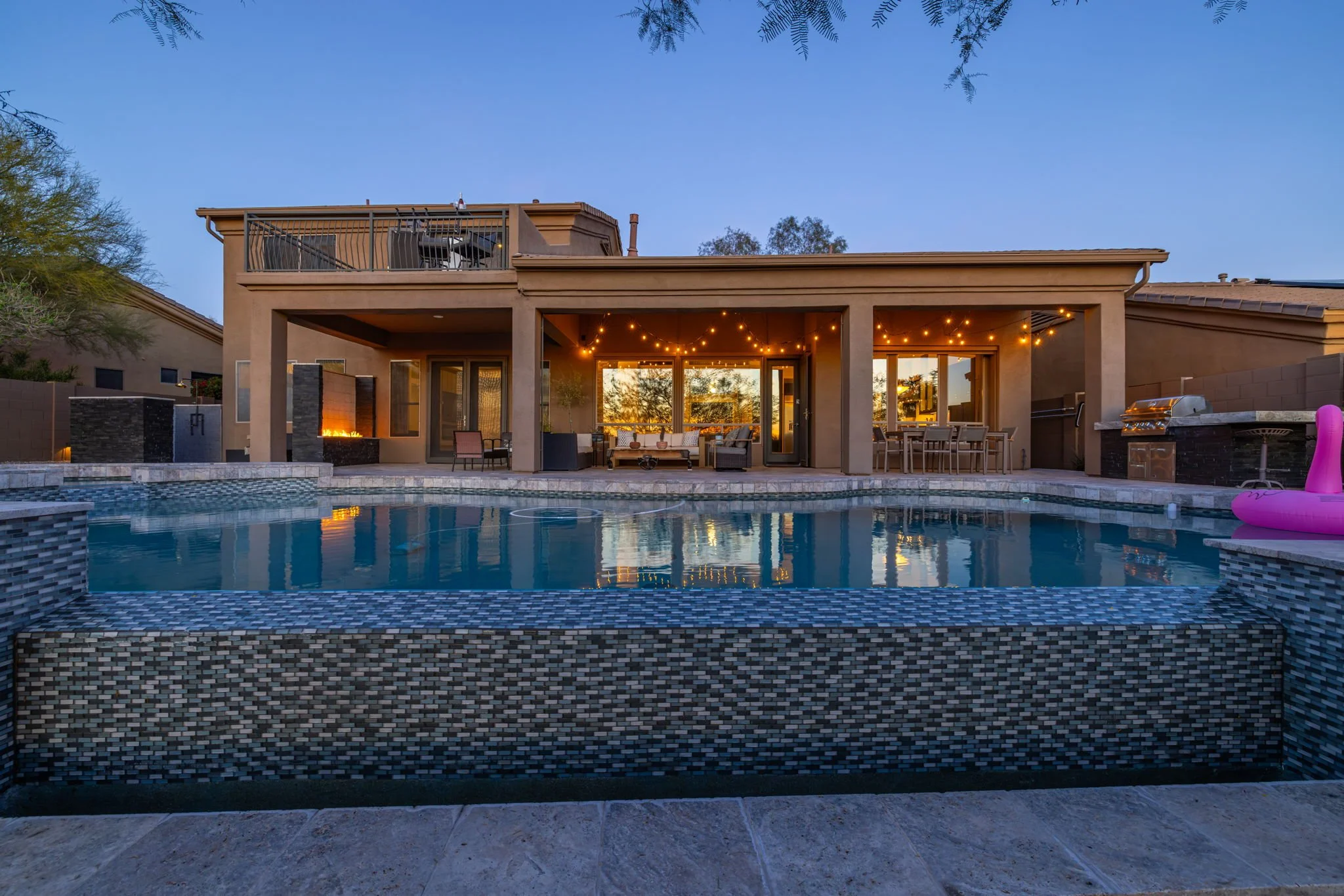 Backyard view of a modern house with a swimming pool, outdoor seating area, string lights, and a second-story balcony at dusk.
