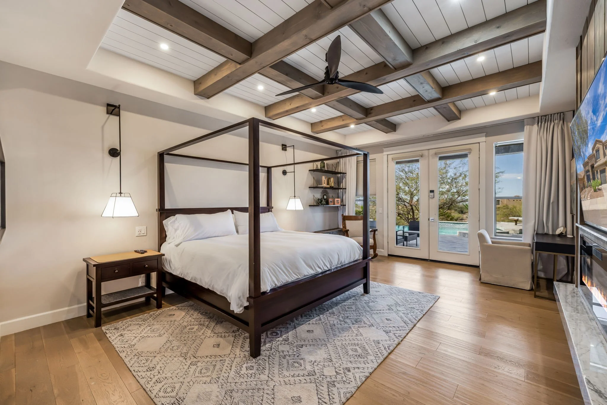 A spacious bedroom with a four-poster bed, side tables, hanging pendant lights, a sitting chair, and a large television. The room features wooden flooring, a patterned rug, exposed wooden ceiling beams, and windows with a view of a pool outside.