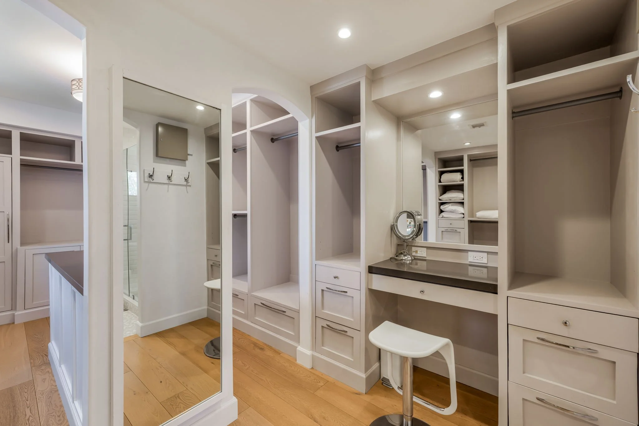 Walk-in closet with built-in shelves, drawers, a mirror, a stool, and a small vanity area. In the background, bathroom with a shower and a small bench.