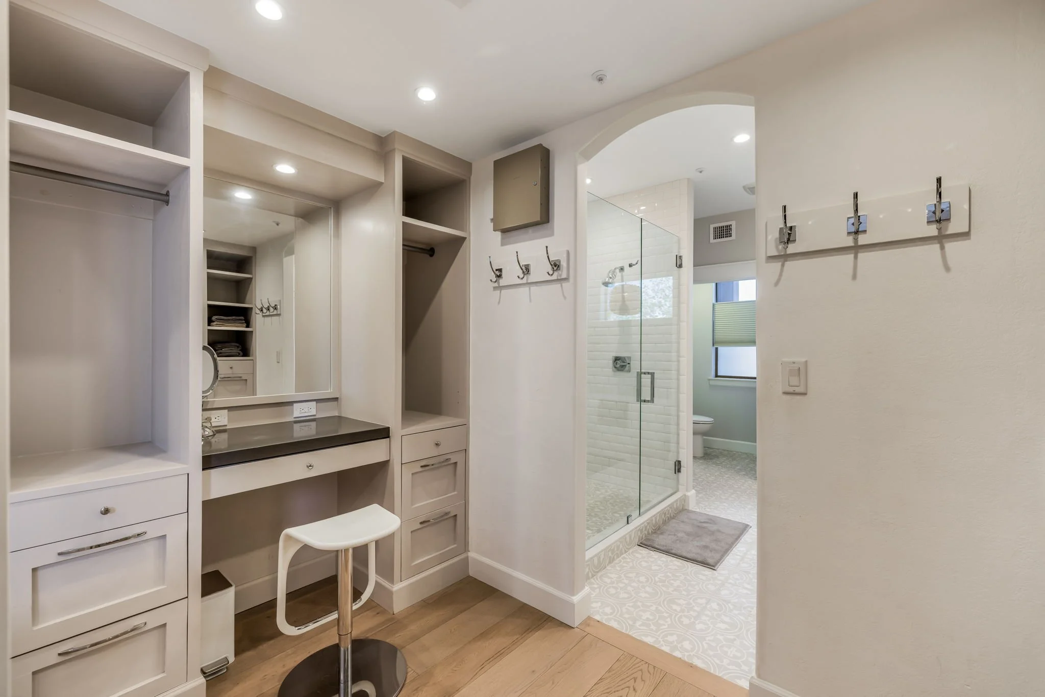 Interior of a room with built-in white shelving, a mirror, a small seating stool, and a bathroom with a glass shower enclosure, a toilet, and a window.